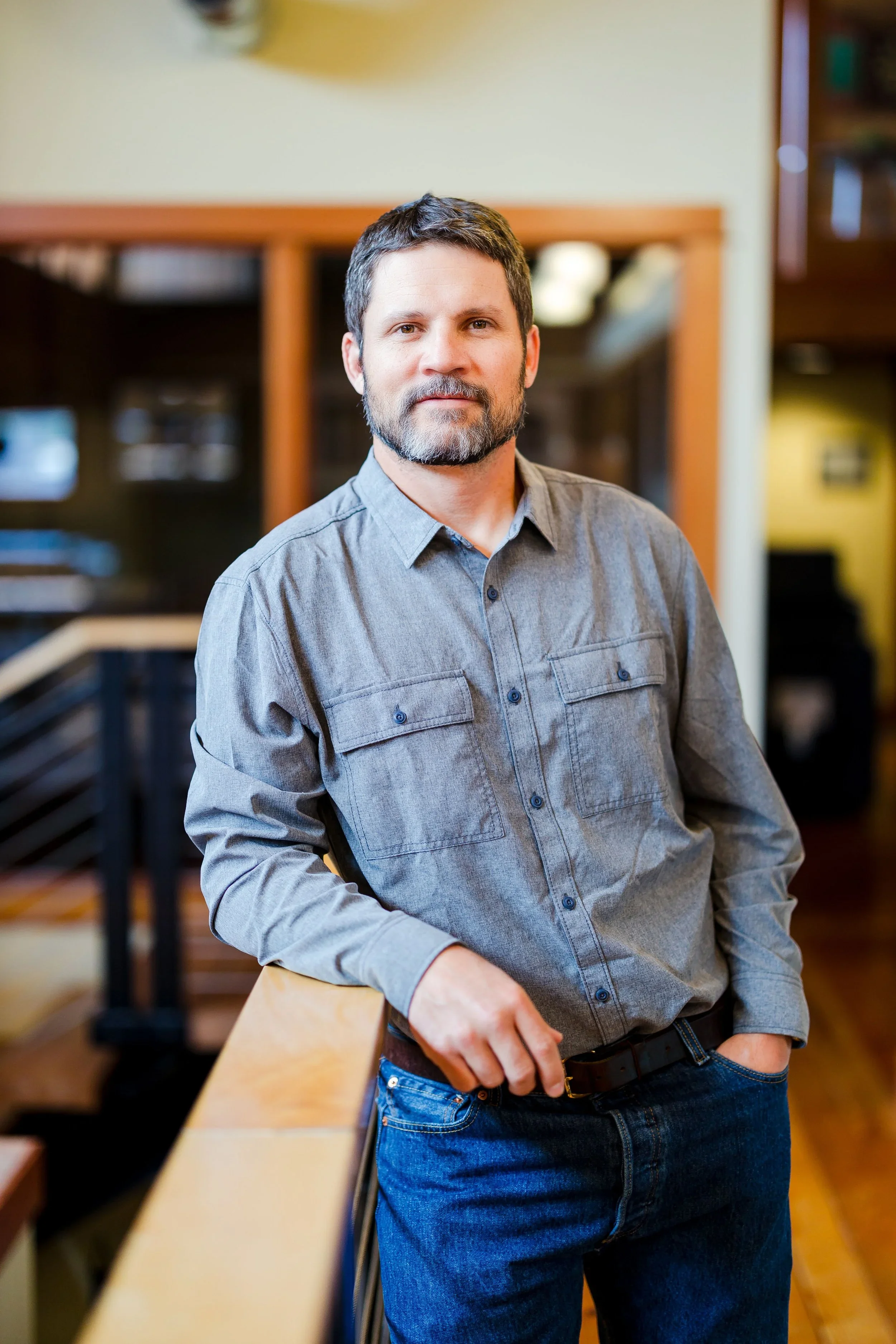 A man with a beard and short dark hair, wearing a gray button-up shirt and jeans, leaning against a railing indoors.
