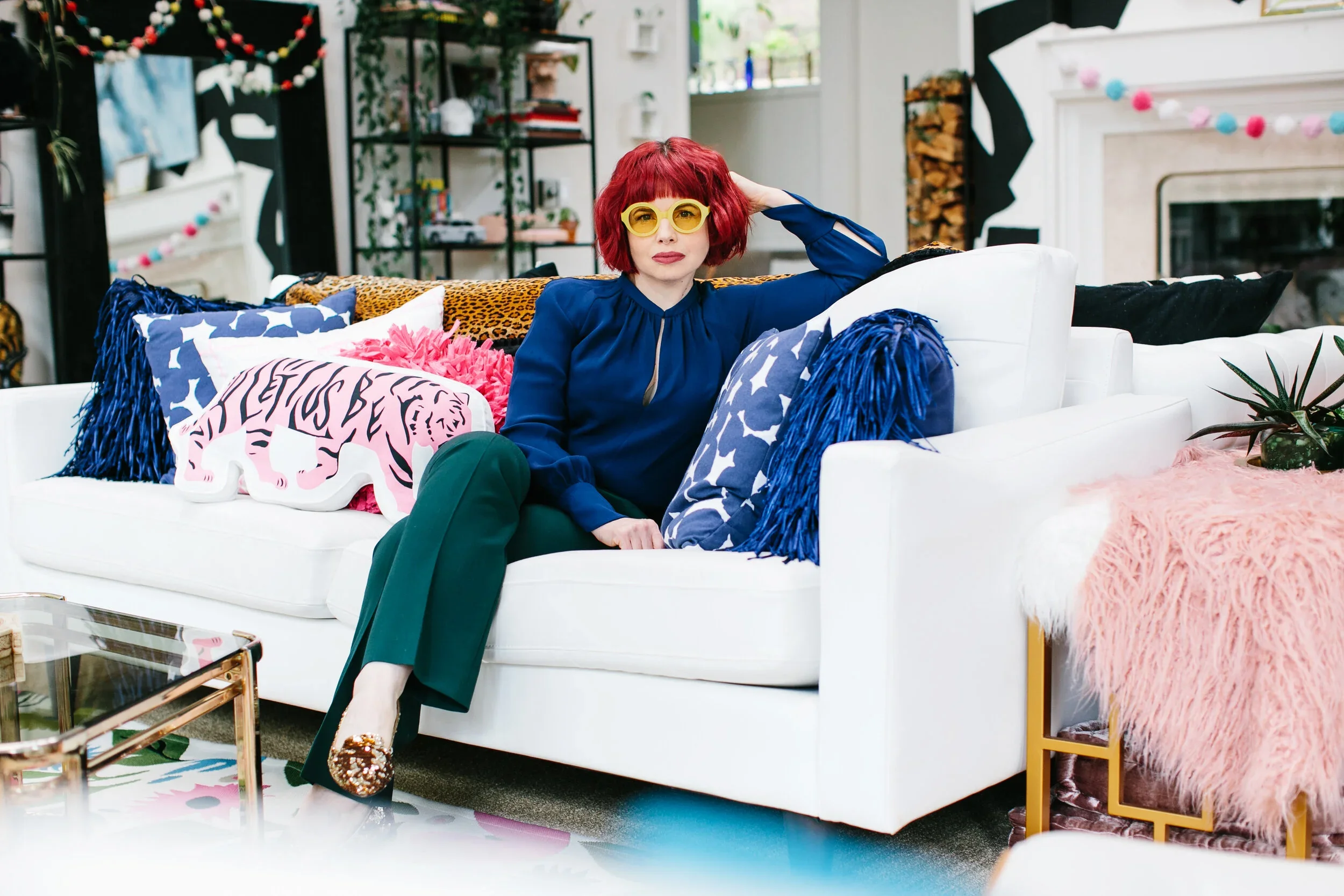 A woman with bright red hair, yellow glasses, and a blue blouse sitting on a white couch with colorful pillows and a pink furry blanket in a decorated living room.