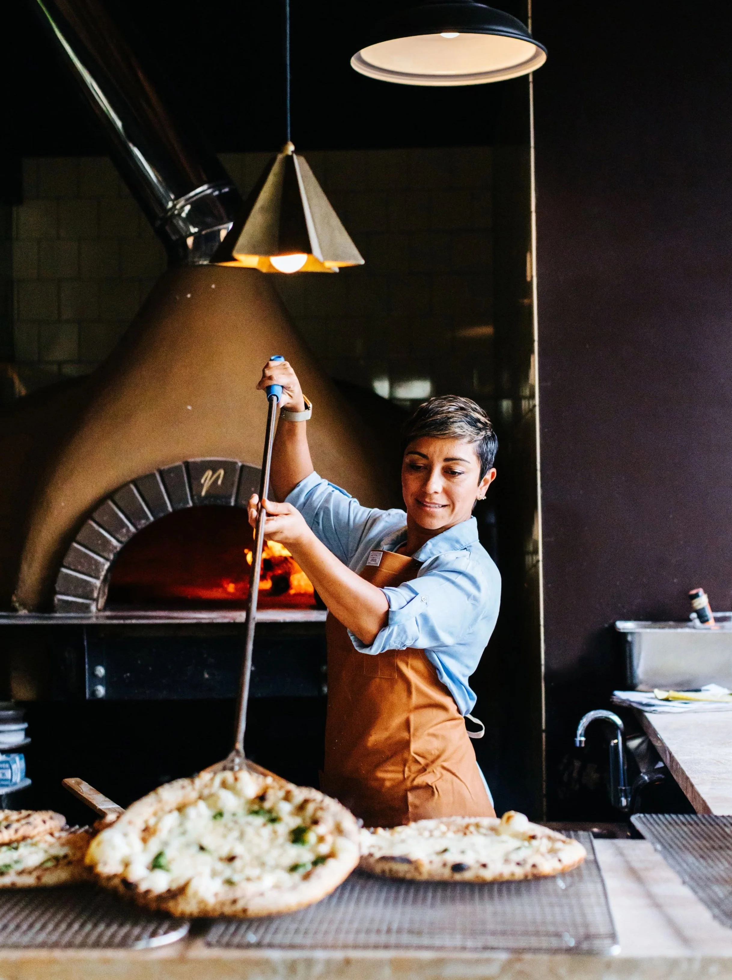 A woman with short dark hair and wearing a blue shirt and brown apron is taking a pizza out of a wood-fired oven in a pizzeria. The scene includes pizzas on a counter in the foreground, with one pizza visible. There is a black wall and a lamp hanging