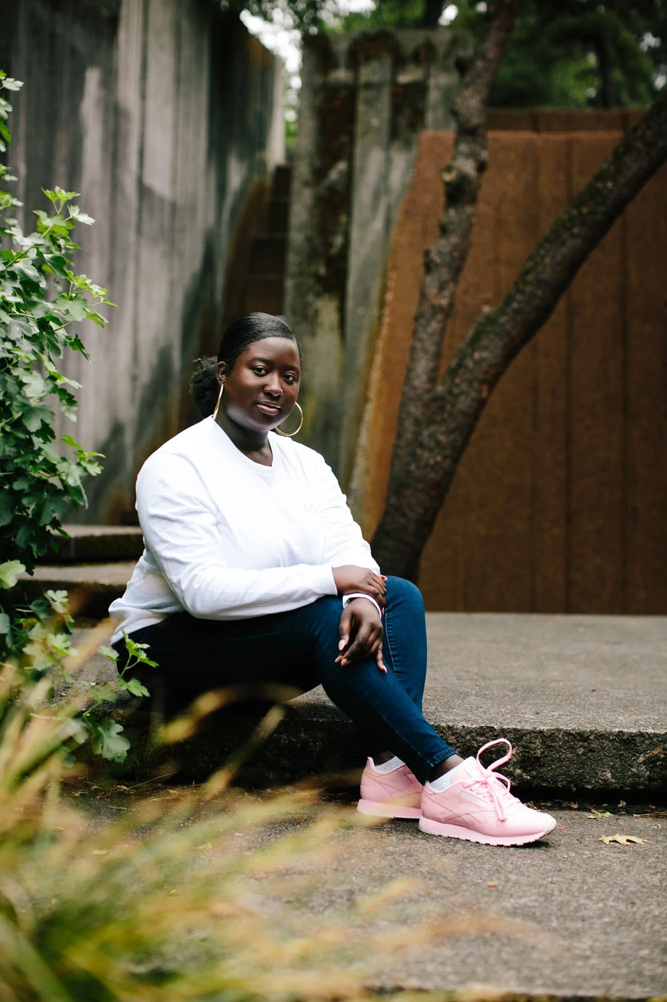 A woman sitting on a concrete step outdoors, wearing a white long-sleeve shirt, dark jeans, and pink sneakers, with greenery and a tree behind her.