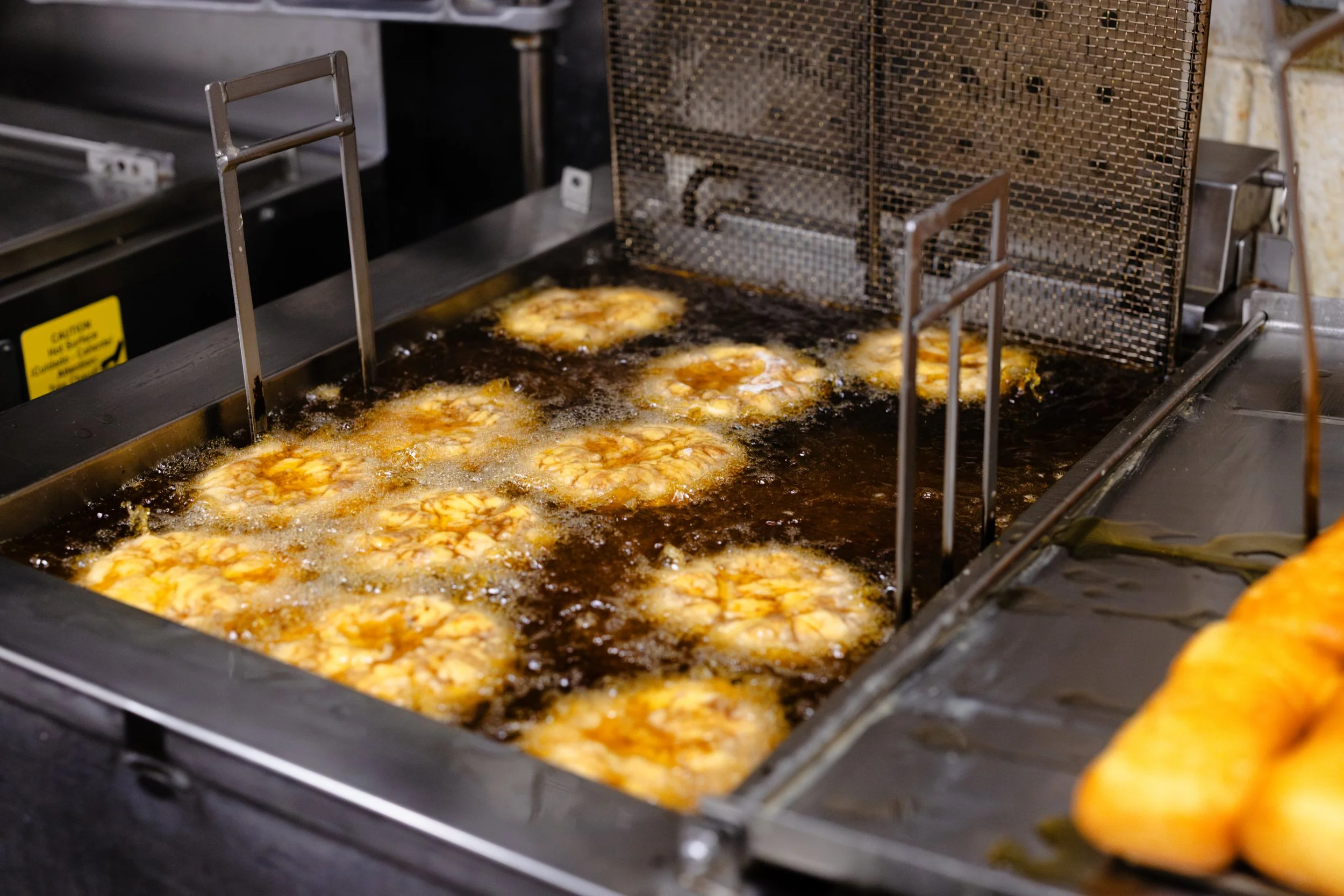 Frying plantains in hot oil on a commercial stove, with a metal mesh screen and yellow caution sign nearby.
