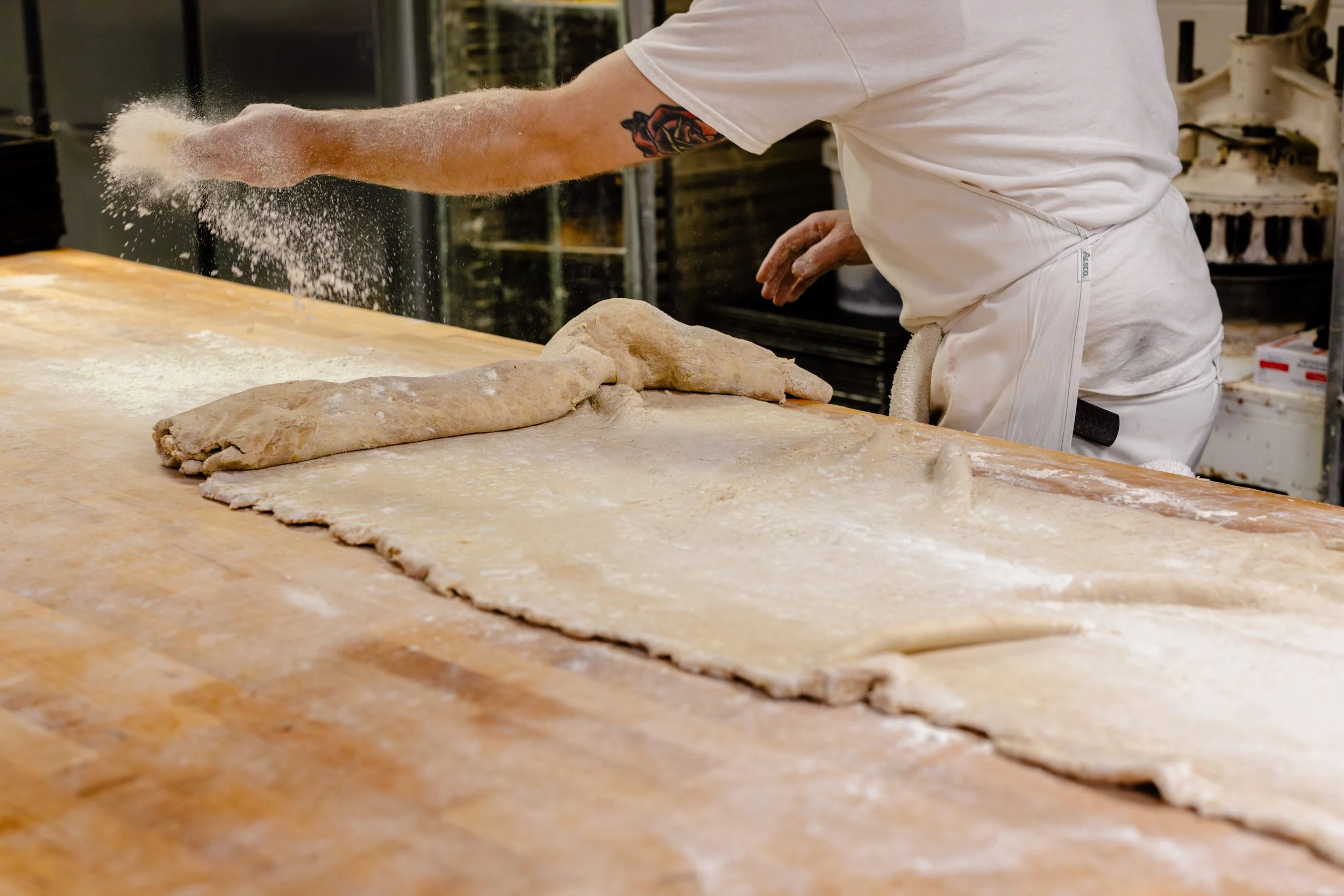 Bakery worker tossing flour in the air while preparing bread dough on a wooden surface.