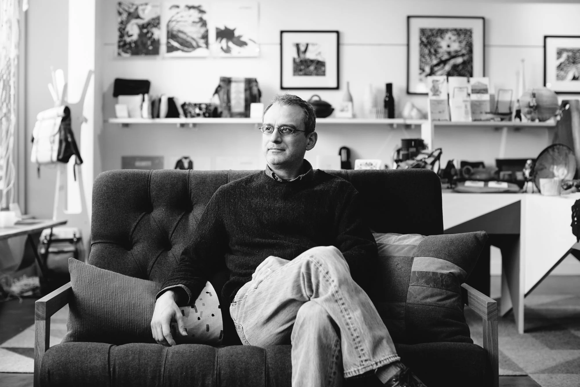 A man sitting on a dark fabric couch in a room with shelves and framed pictures on the wall behind him. He wears glasses, a dark sweater, and light-colored jeans, with one arm resting on a cushion.