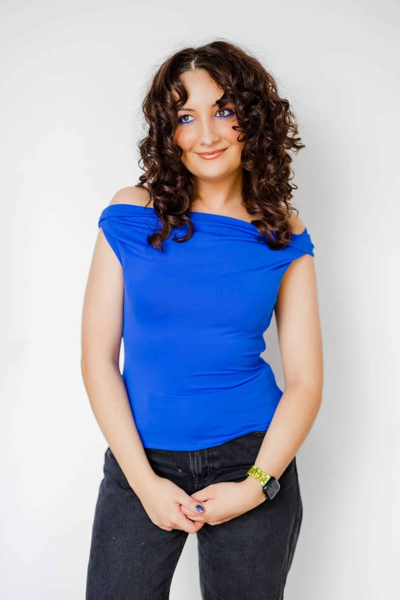 A woman with curly brown hair wearing a blue off-the-shoulder top, black pants, and a smartwatch, standing against a plain white background.