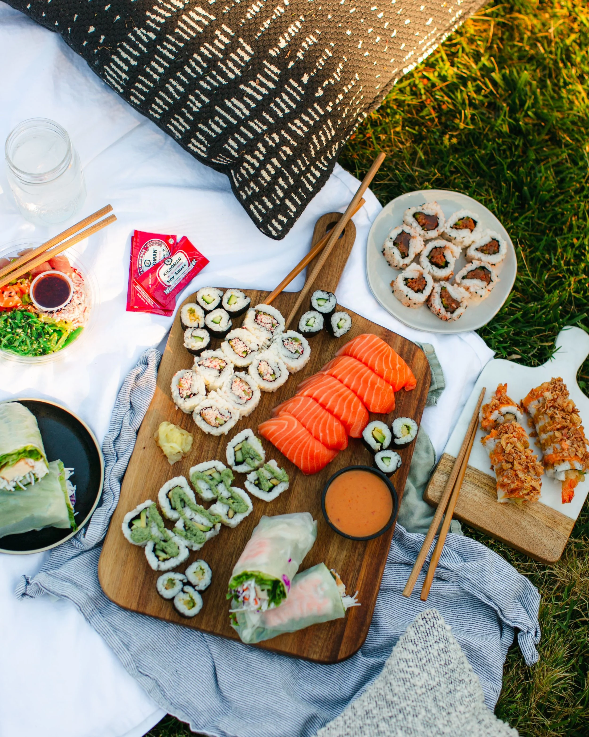 A variety of sushi dishes on a wooden cutting board, with salmon, rolls, and sushi wraps, surrounded by plates of sushi, dipping sauces, and beverages on a picnic setup outdoors.