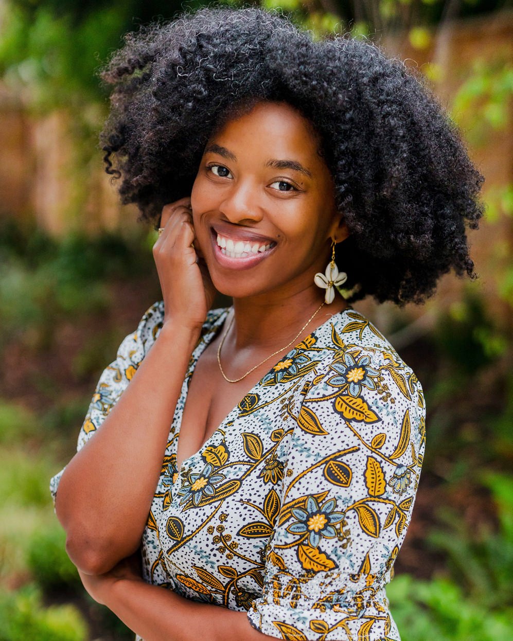 A woman with natural curly hair smiling outdoors, wearing a patterned dress with flowers and leaves, and floral earrings.