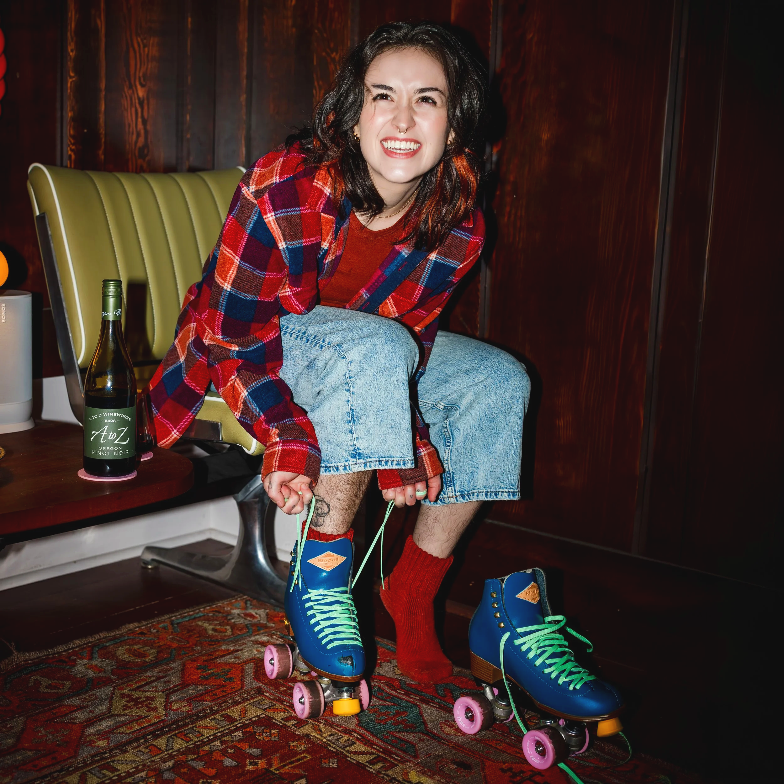 A person with dark, curly hair smiling while sitting on a skateboard. They are wearing a red plaid shirt over a red top, denim shorts, red socks, and blue roller skates. The background features a wooden wall and a patterned rug on the floor.
