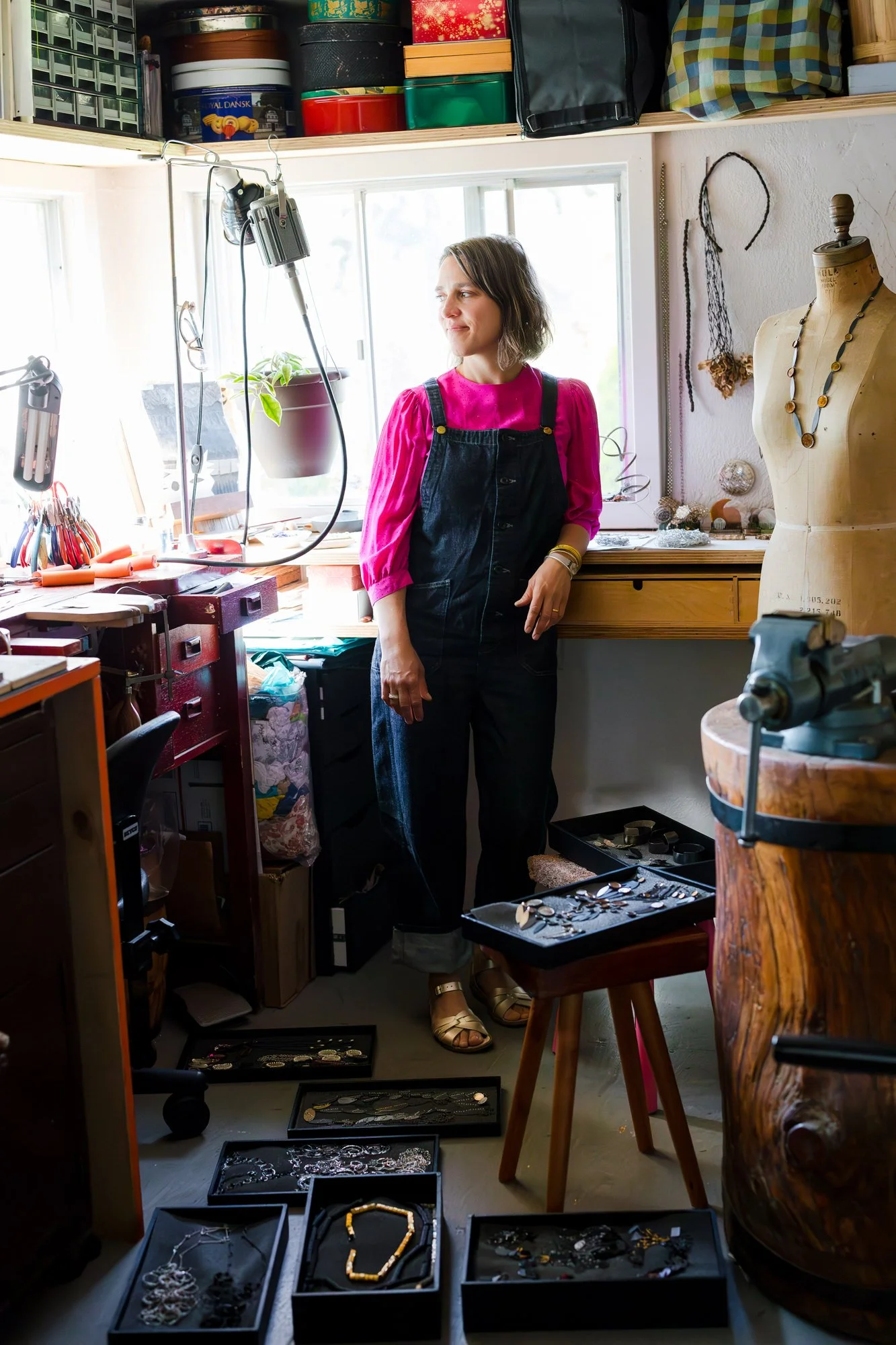 A woman in pink and black overalls stands in a jewelry workshop, surrounded by jewelry and tools, with a mannequin wearing a necklace nearby and a window behind her.
