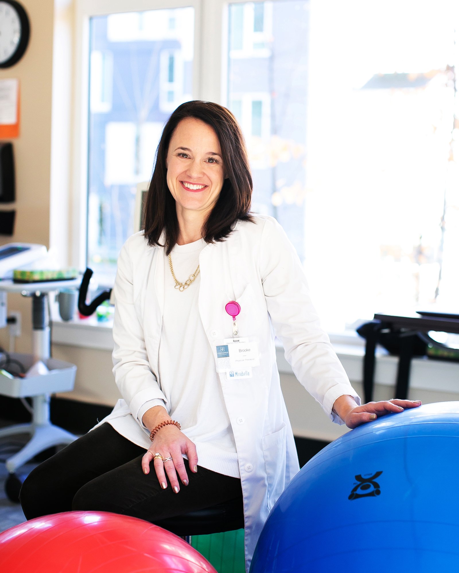 A smiling female healthcare professional in a white lab coat sitting in a medical office with exercise balls.
