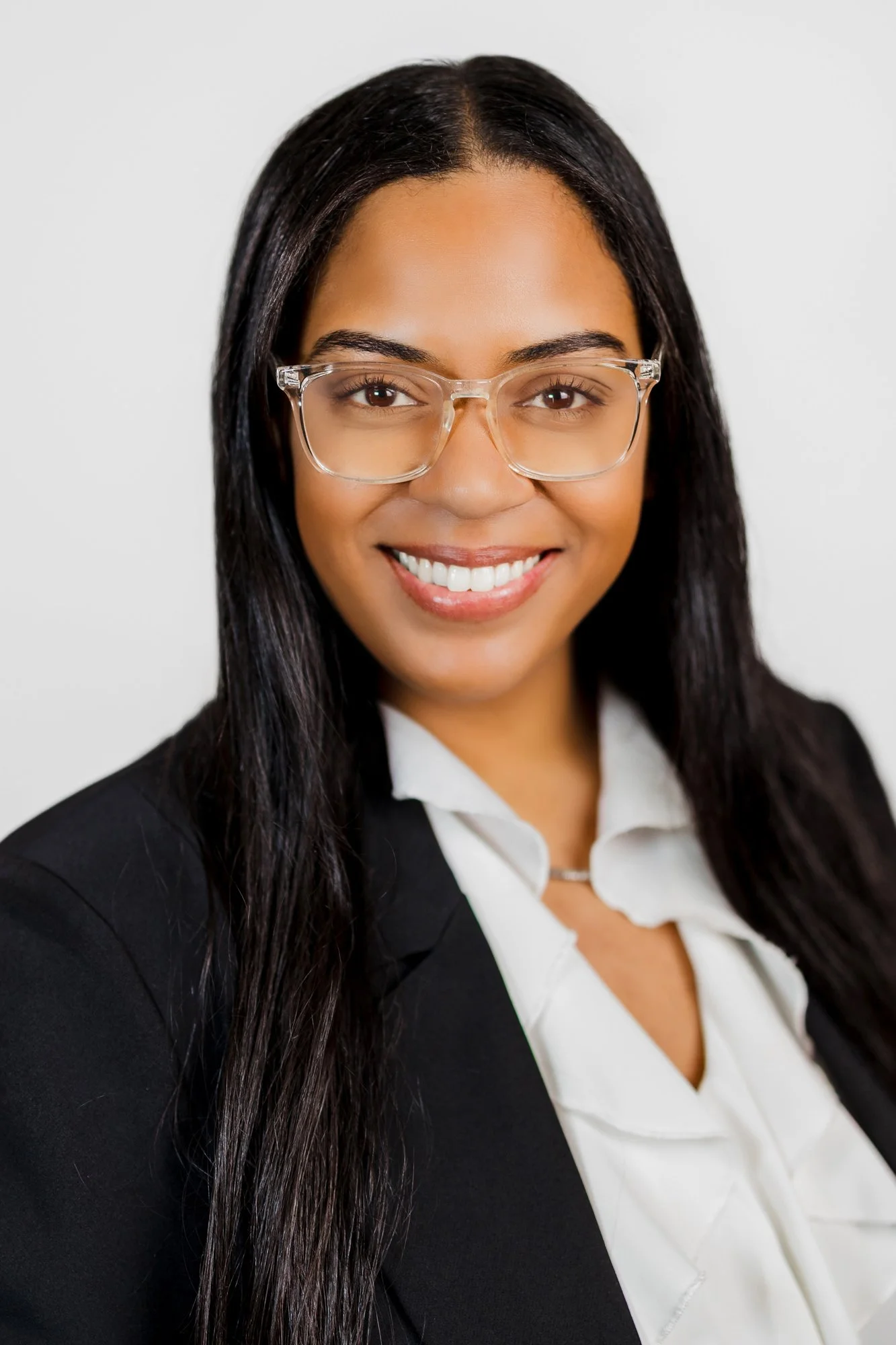 A woman with long dark hair, wearing clear glasses, a white blouse, and a black blazer, smiling against a plain light background.