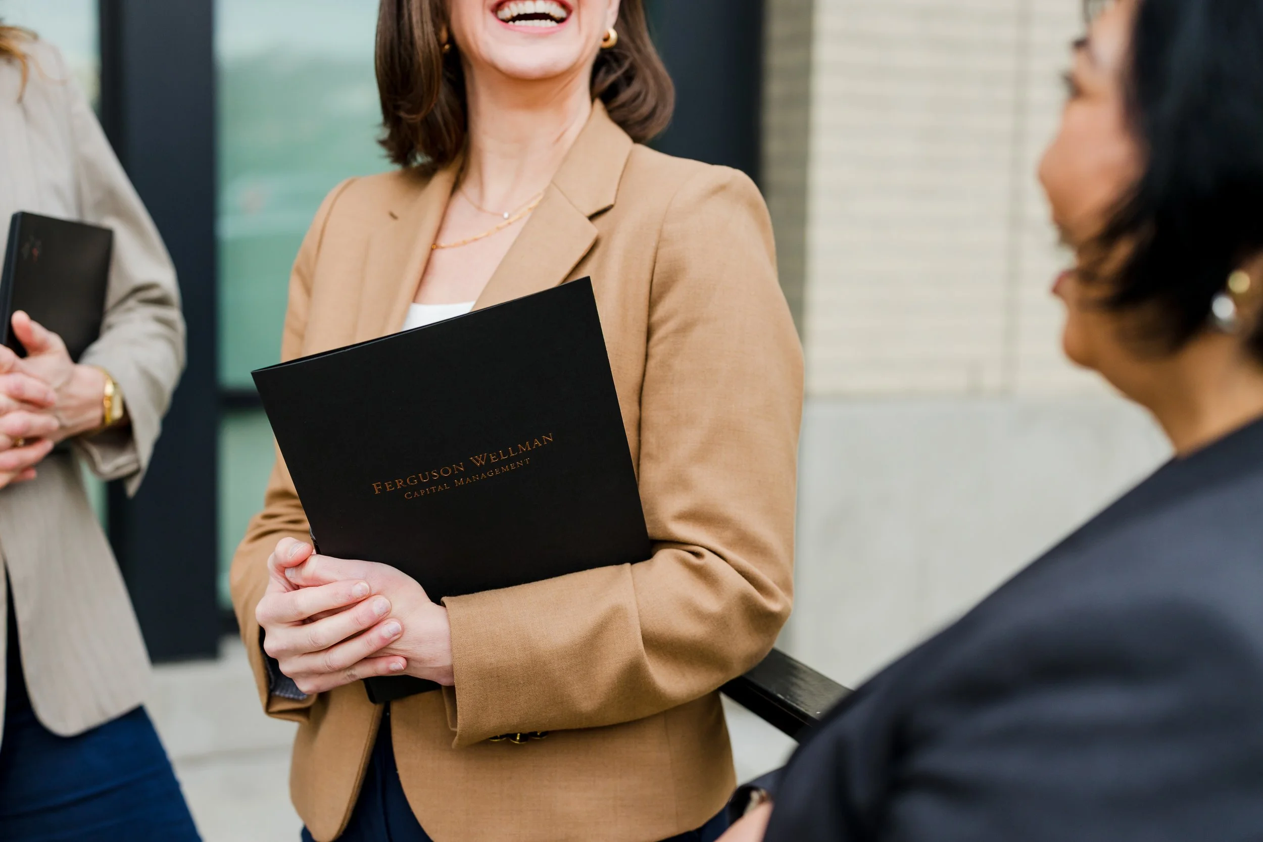 Two women in business attire having a conversation outdoors, one woman is holding a black folder with the words 'Ferguson Wellman Capital Management' on it, and the other woman is holding a notebook.