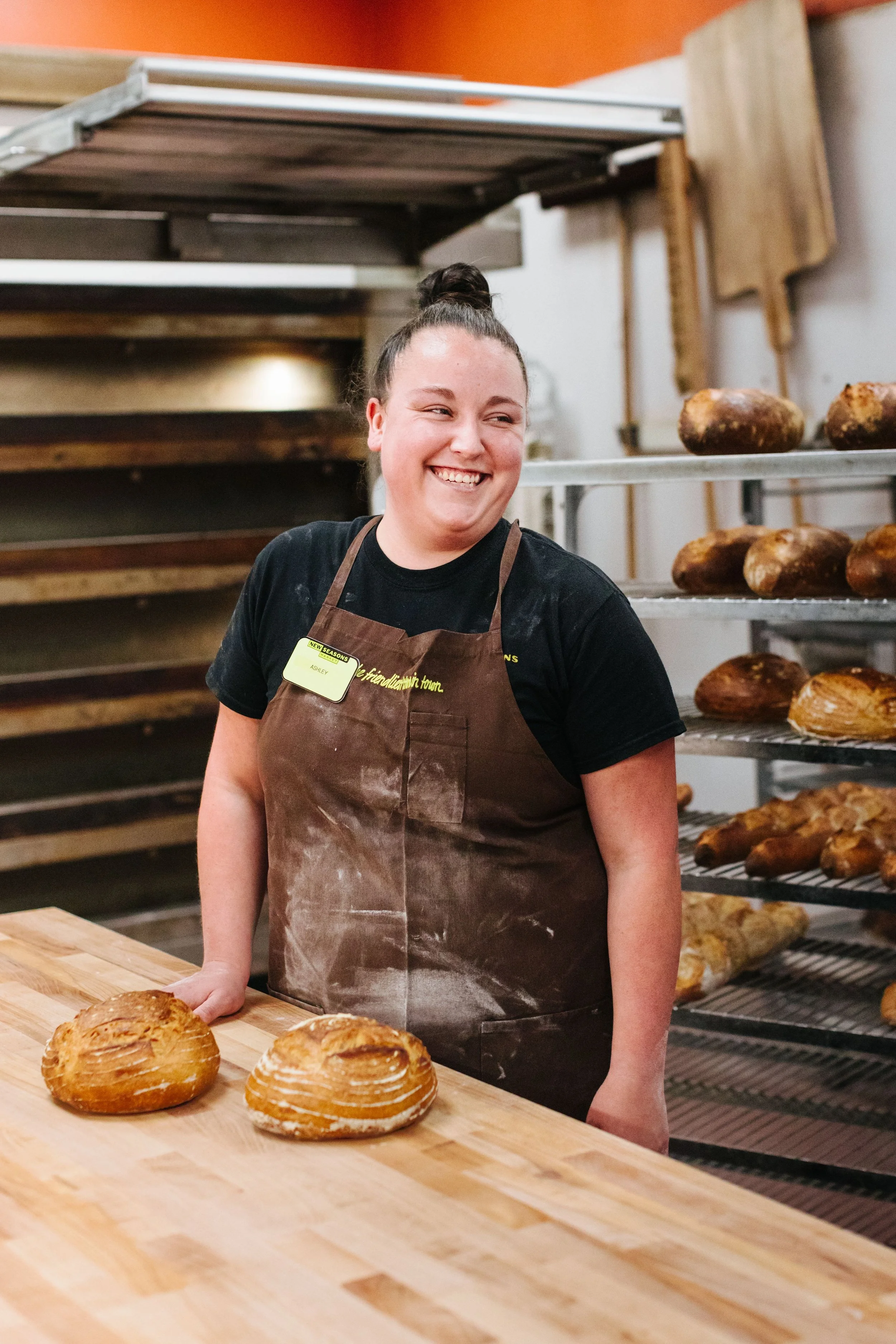 Smiling woman in apron standing behind a wooden counter with two loaves of bread, baking racks of bread in the background.