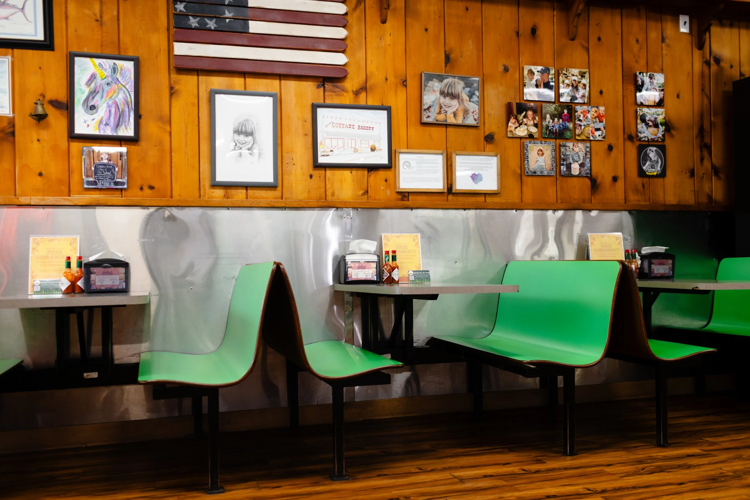 Empty restaurant booth with green seats, wooden wall decorated with framed pictures and an American flag made from wood planks, restaurant condiments on tables.
