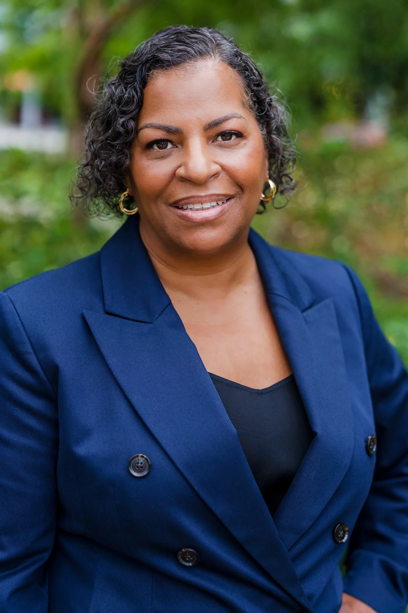 A woman with short curly hair wearing a navy blue blazer and gold hoop earrings outdoors with blurred green trees in the background.