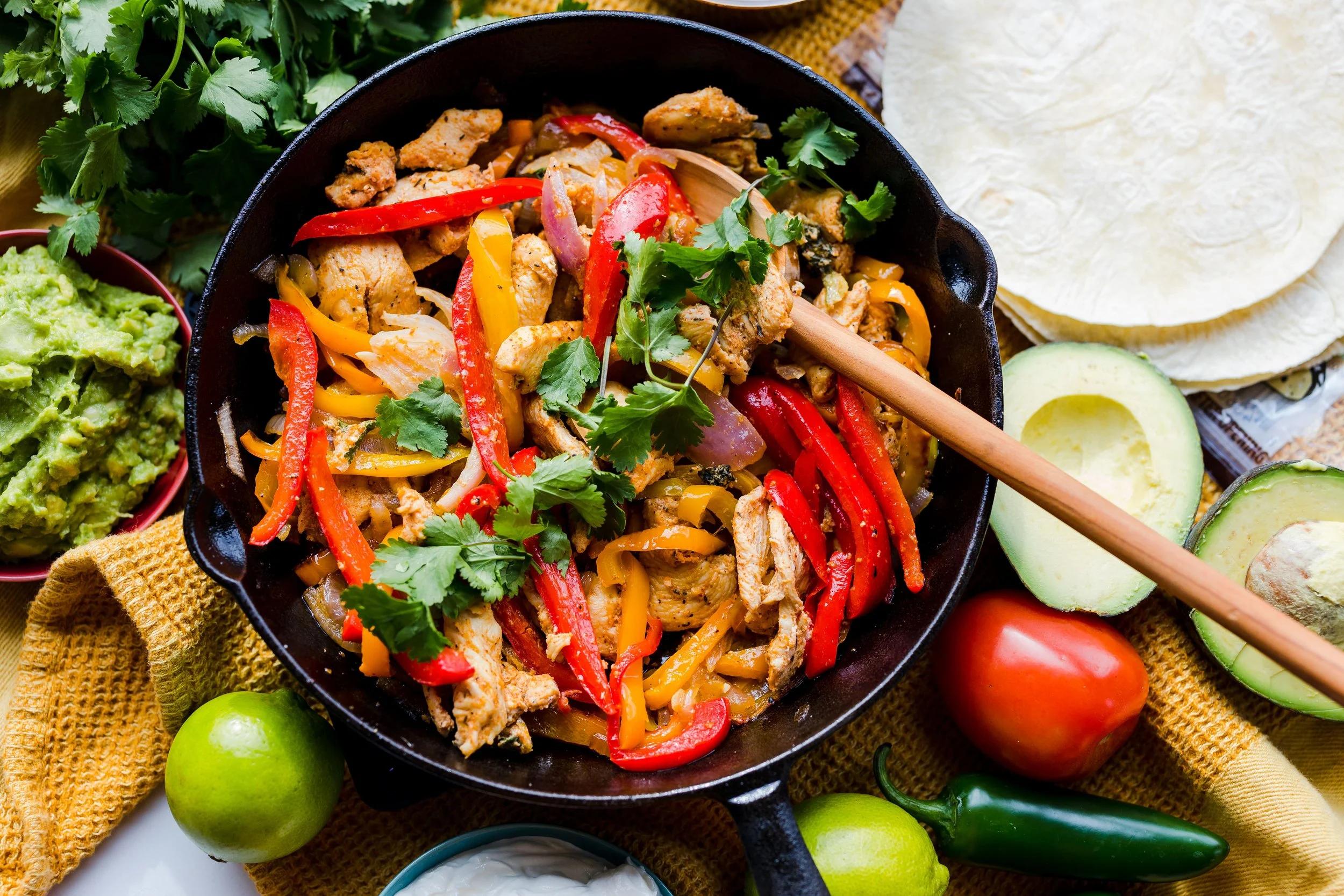 A cast iron skillet filled with chicken fajitas, red and yellow bell peppers, onions, and garnished with cilantro, surrounded by avocado slices, tomatoes, corn tortillas, guacamole, sour cream, and jalapeño peppers on a yellow cloth.