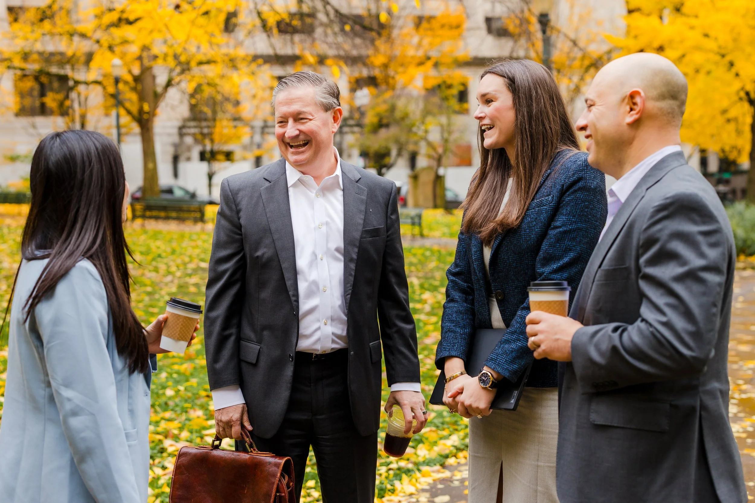 Four professionally dressed people, two men and two women, are talking and laughing outdoors in a park with fall foliage. They are holding coffee cups and a briefcase.