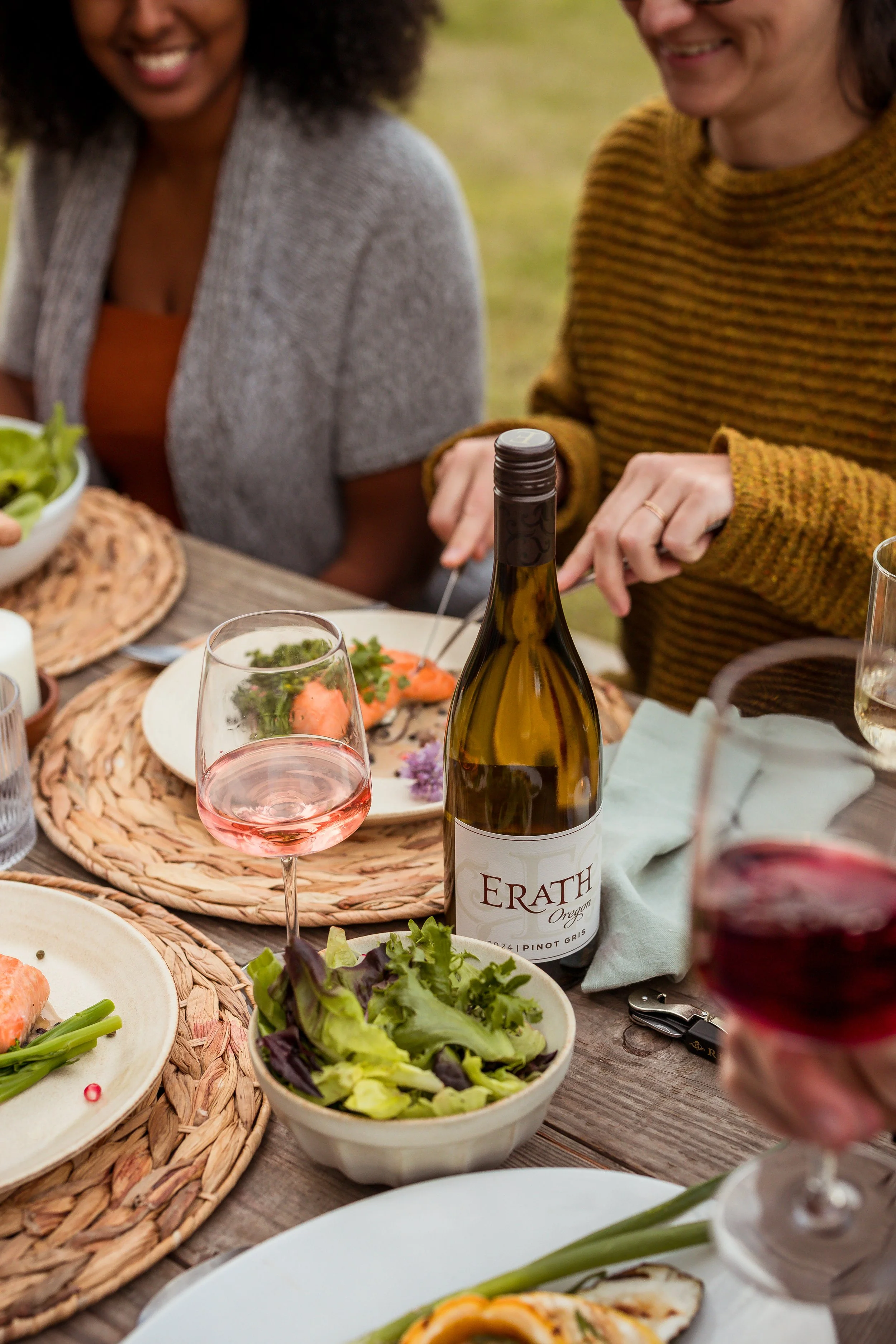 People enjoying a meal outdoors, with a table set with wine glasses, a bottle of wine labeled 'Erath Oregon Pinot Gris', salads, and plates of food.