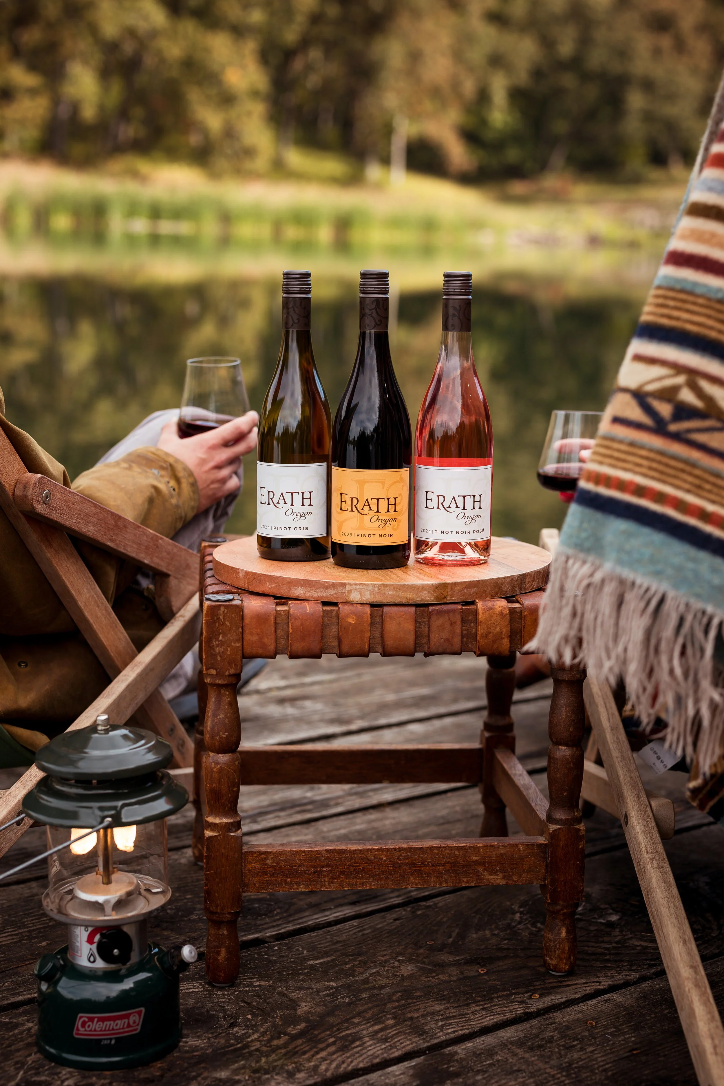 Three bottles of Erath Oregon wine on a small wooden table beside two people enjoying glasses of wine outdoors near a lake, with trees in the background.