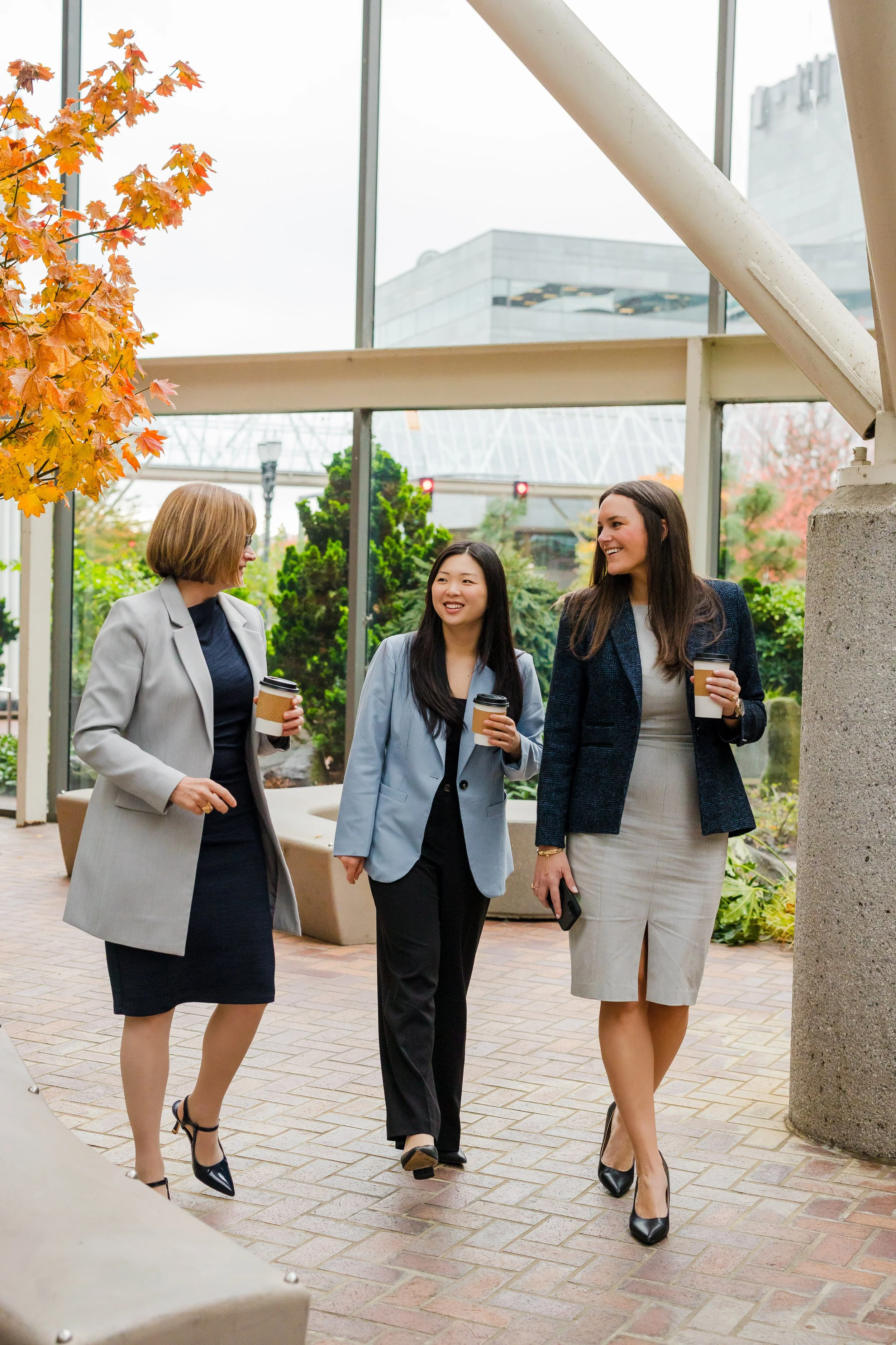 Three women in business attire walking and talking outdoors, each holding a coffee cup.