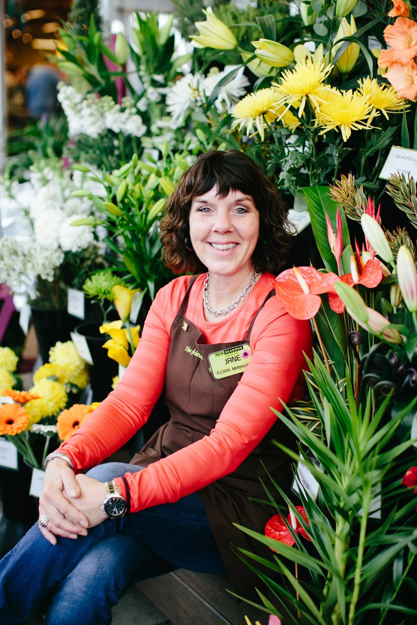 A woman with shoulder-length dark brown hair, smiling, sitting in front of various colorful flowers in a flower shop. She is wearing a red long-sleeve shirt, a brown apron with a name tag that reads "Jane, Floral Manager," a silver chain necklace, an