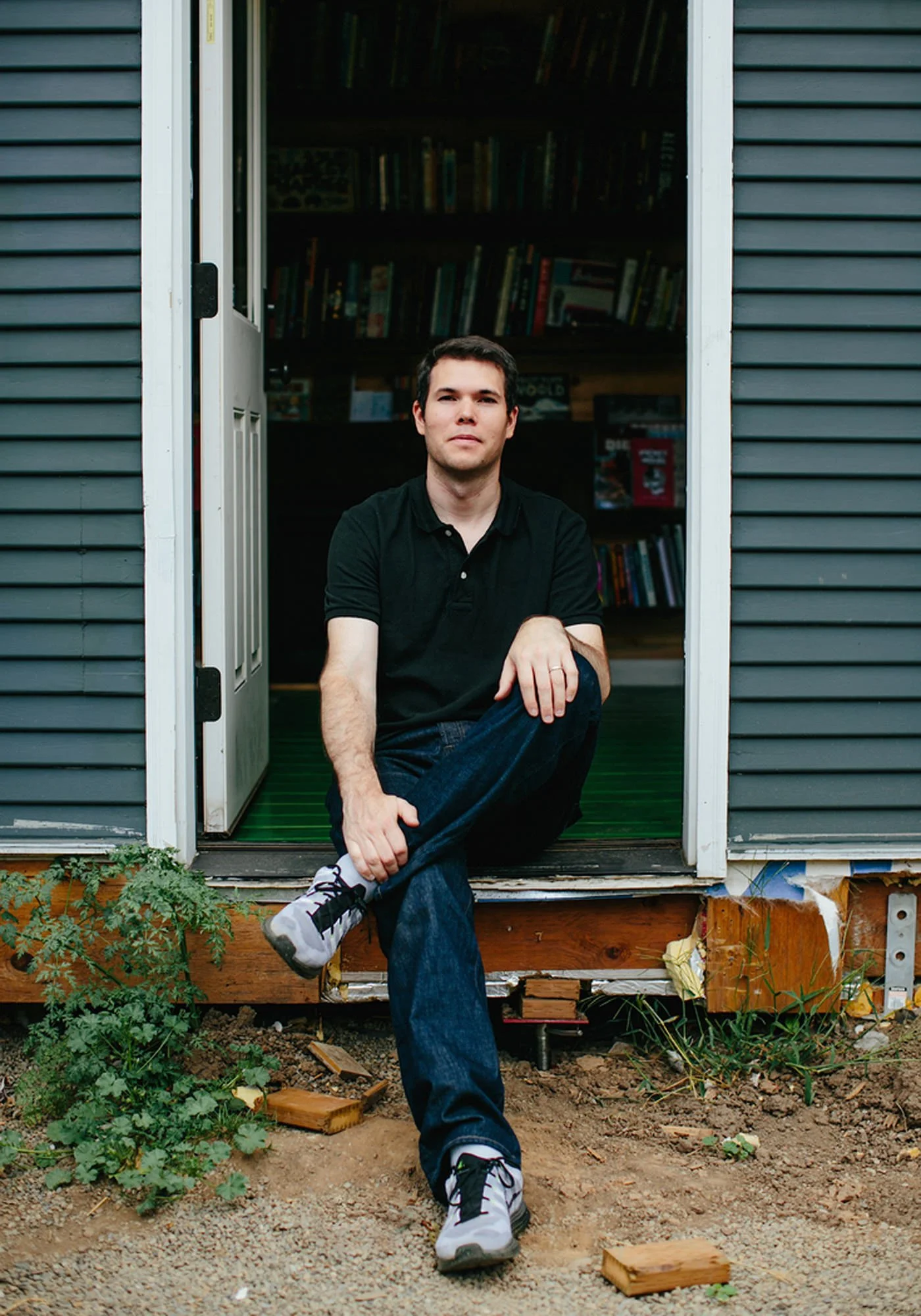 A young man in a black polo shirt and jeans sits on the porch of a house with a bookshelf inside. He is outdoors, with some plants and wooden debris in the foreground.