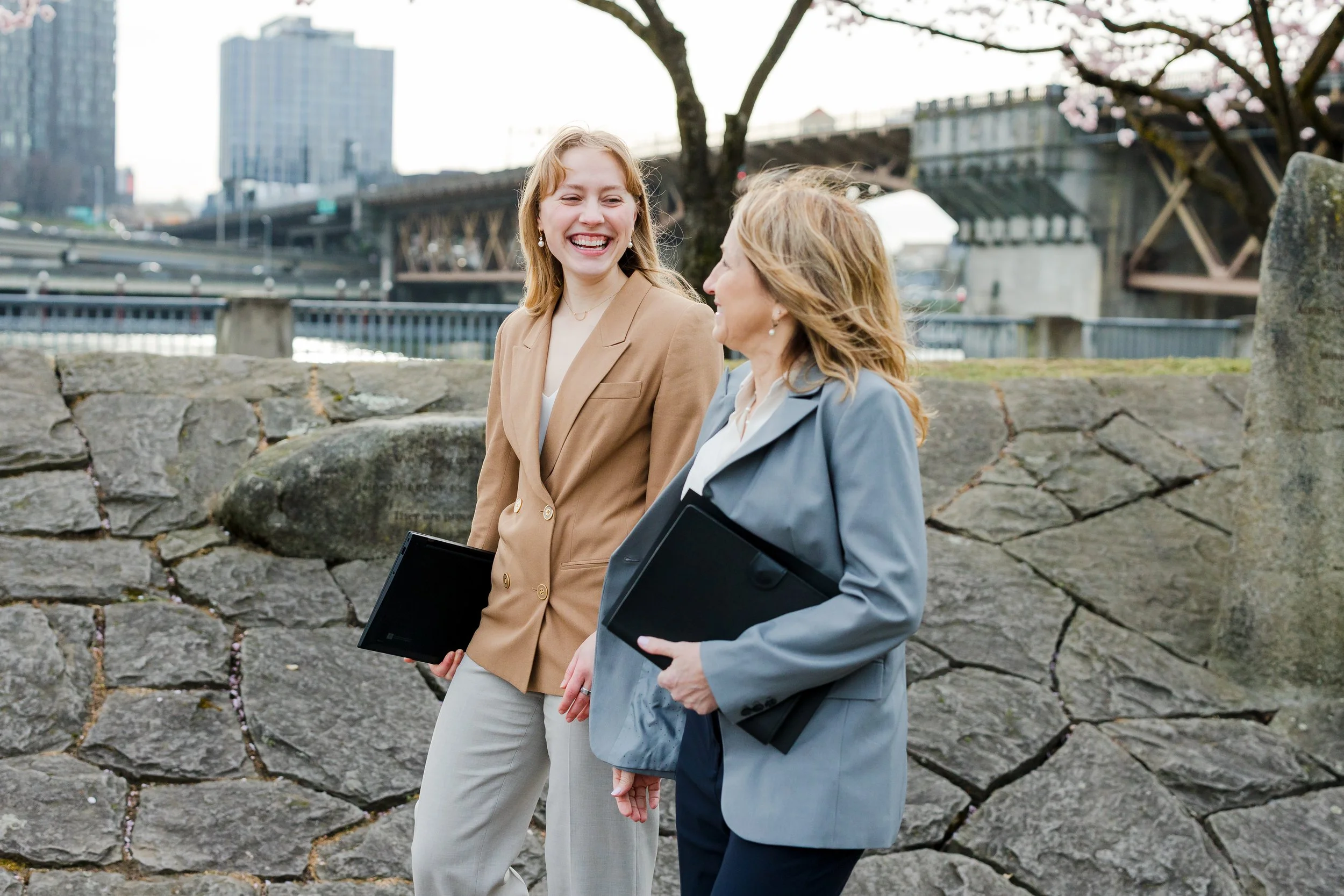 Two women in business attire walking and talking outdoors near a stone wall with city buildings and bridges in the background, holding black folders.