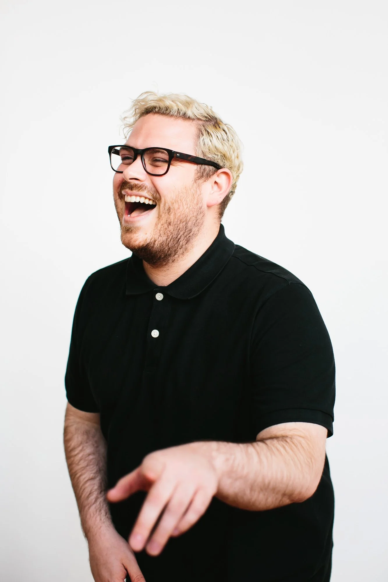 A man with light blonde hair, glasses, and a beard wearing a black polo shirt, laughing with his mouth open and pointing.