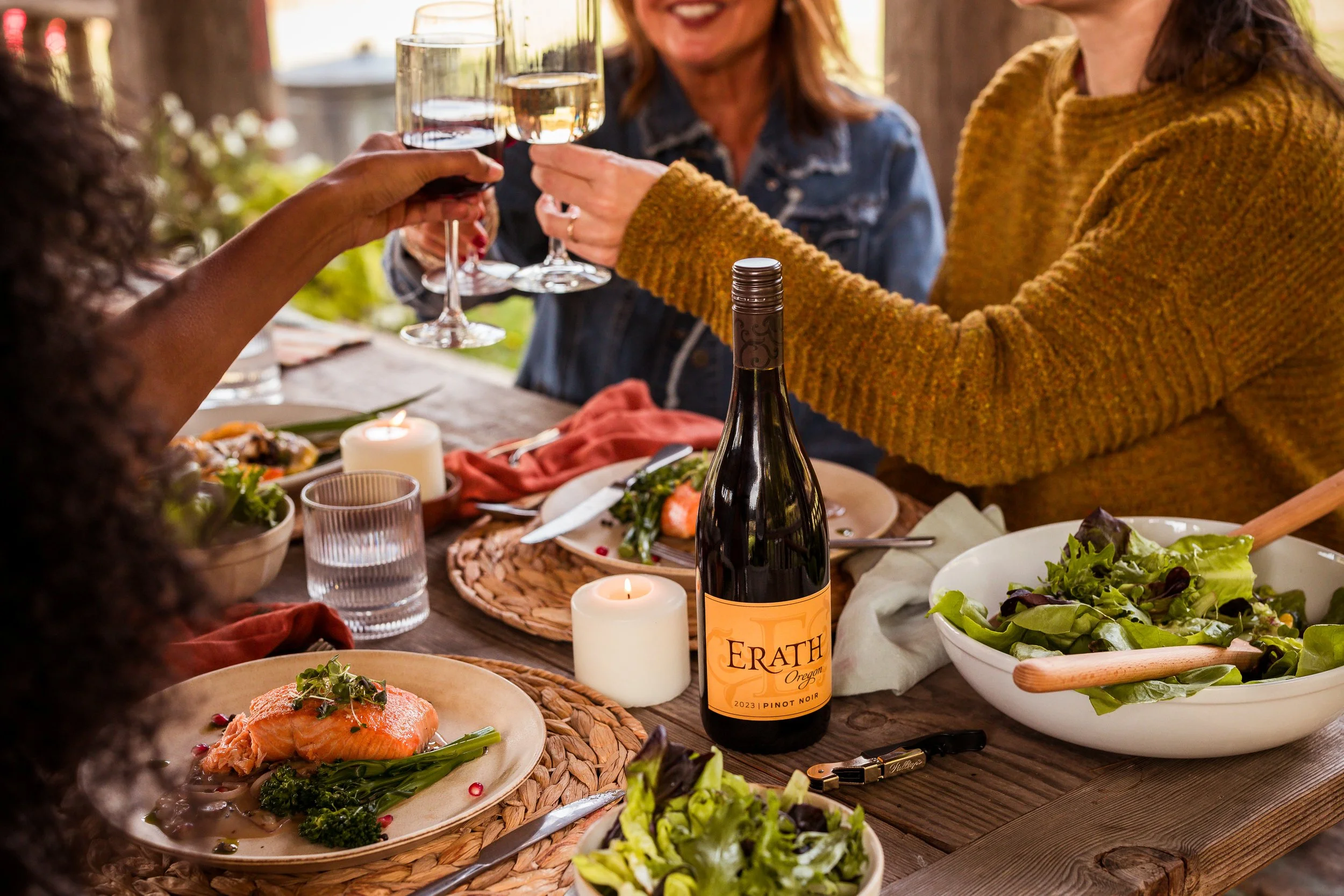 Four women celebrating at a dinner table, clinking wine glasses, with a bottle of wine, salad, and salmon dish on the table.