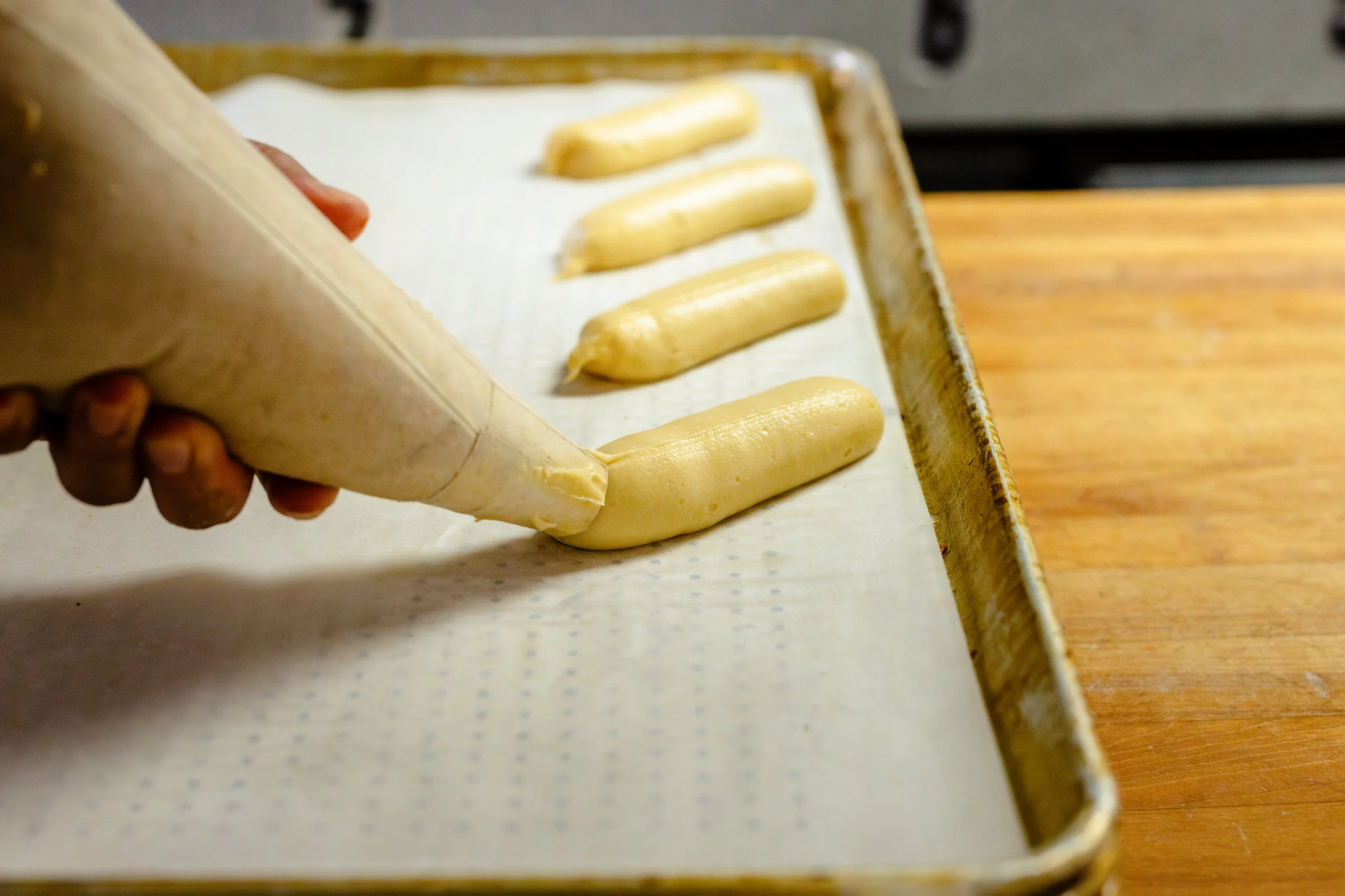 A person piping batter onto a baking sheet lined with parchment paper, with several piped dollops in a row.