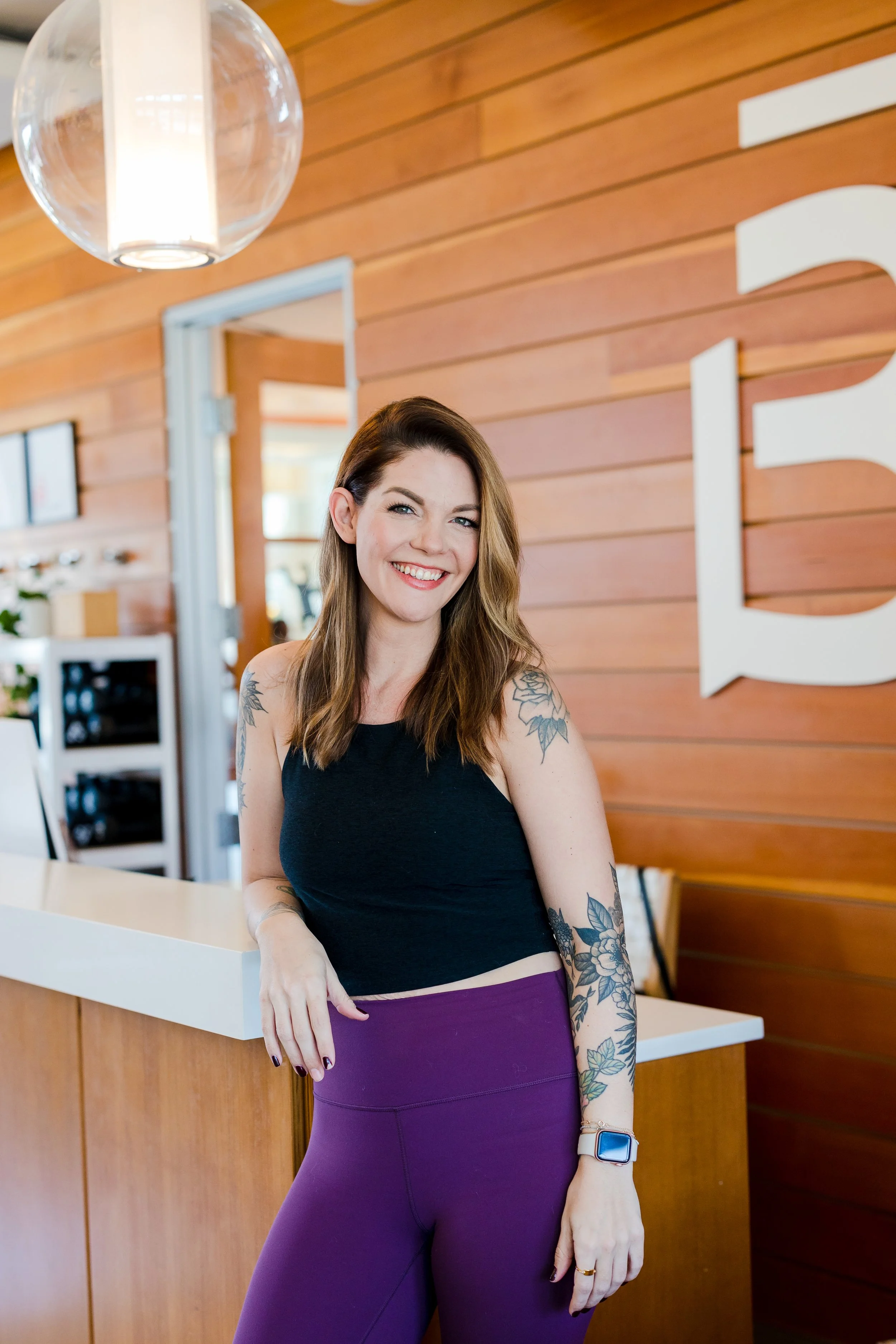 A smiling woman with brown hair and tattoos on her arms, wearing a black sleeveless top and purple leggings, standing at a reception counter in a modern, wood-paneled space.