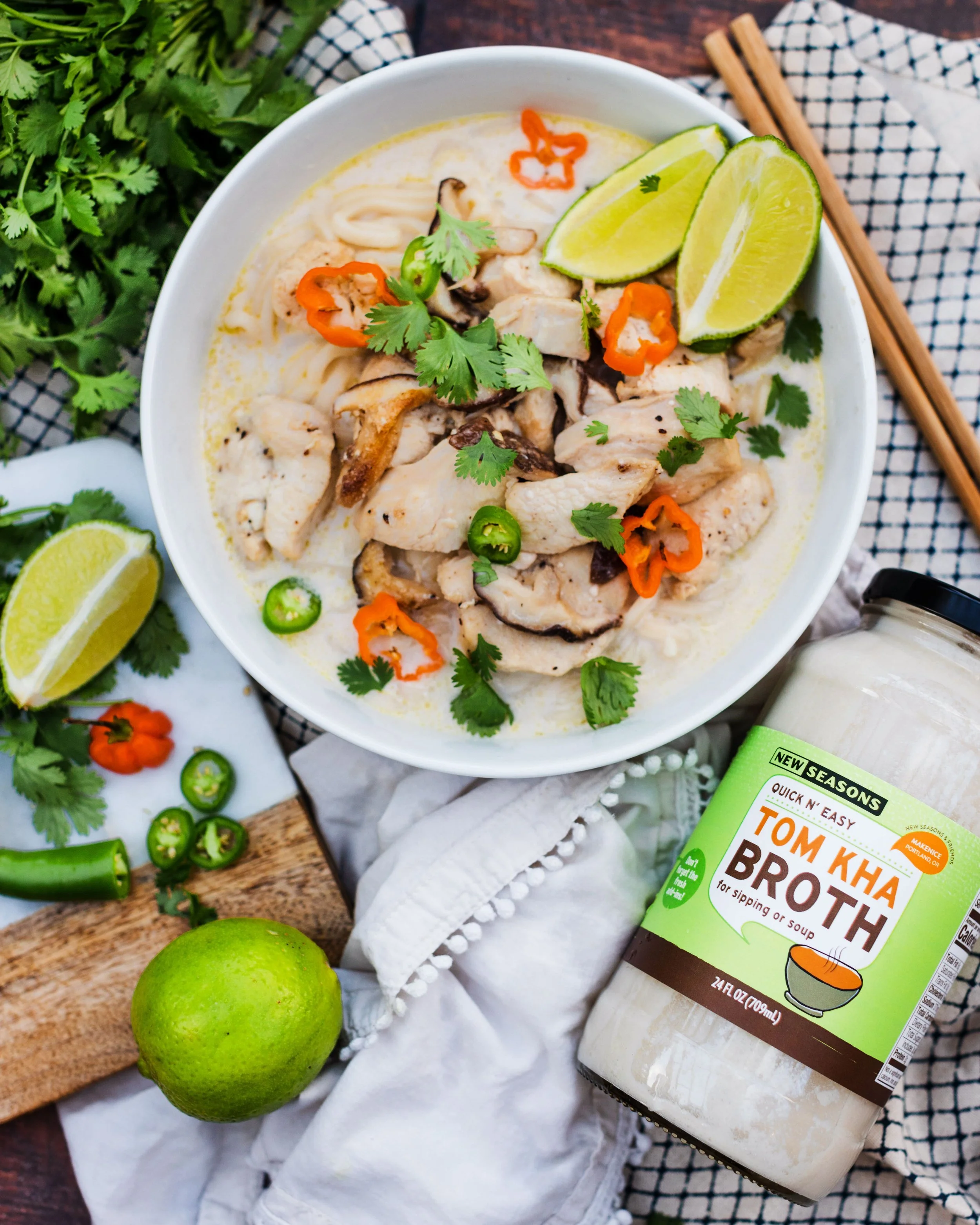 A bowl of chicken and mushroom soup garnished with cilantro, chili peppers, and lime wedges, with fresh cilantro, lime, and chili peppers around it, and a jar of Tom Kha broth nearby.