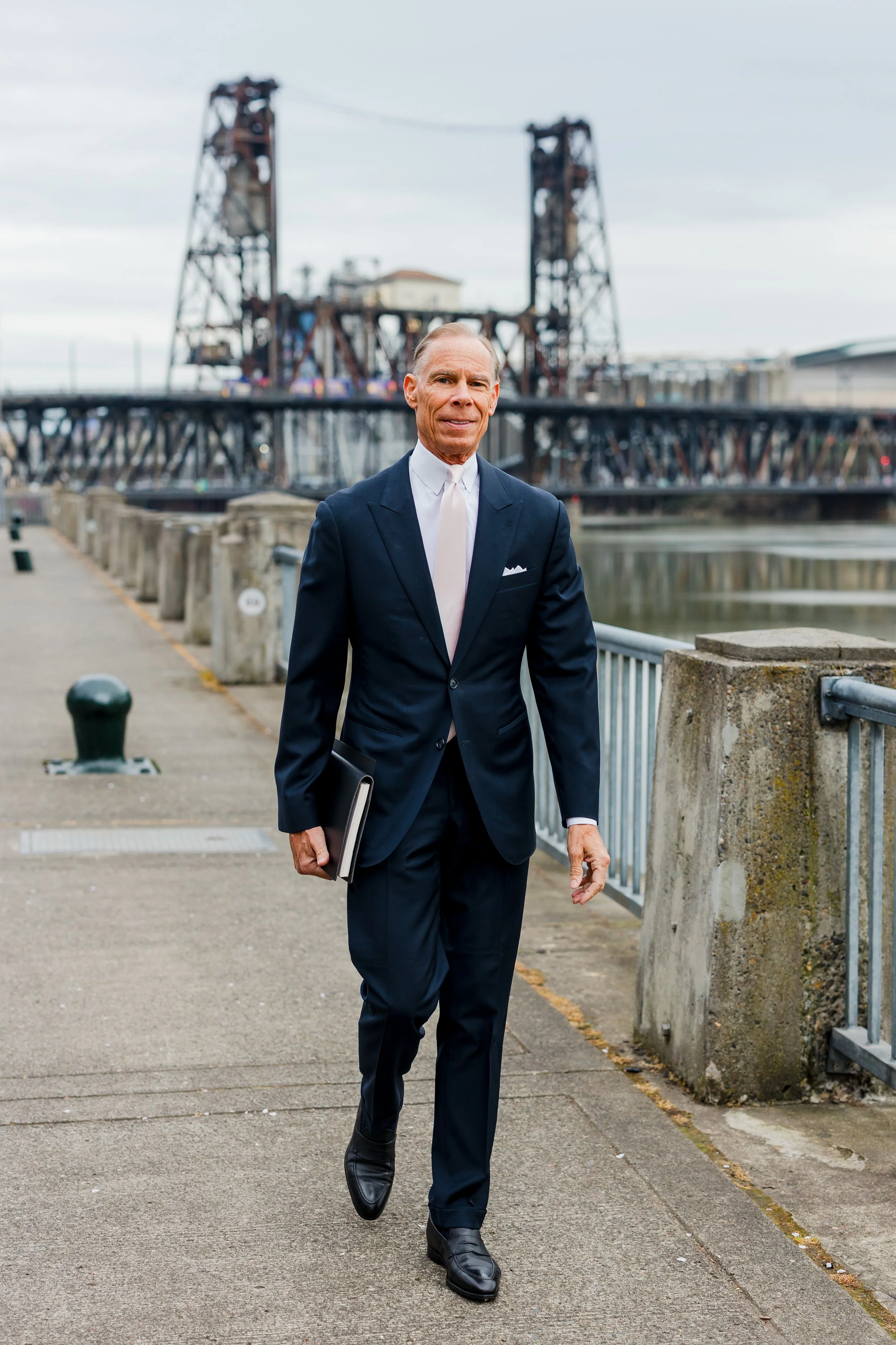 An older man in a navy suit and white shirt walking along a waterfront walkway with industrial structures and a body of water in the background.