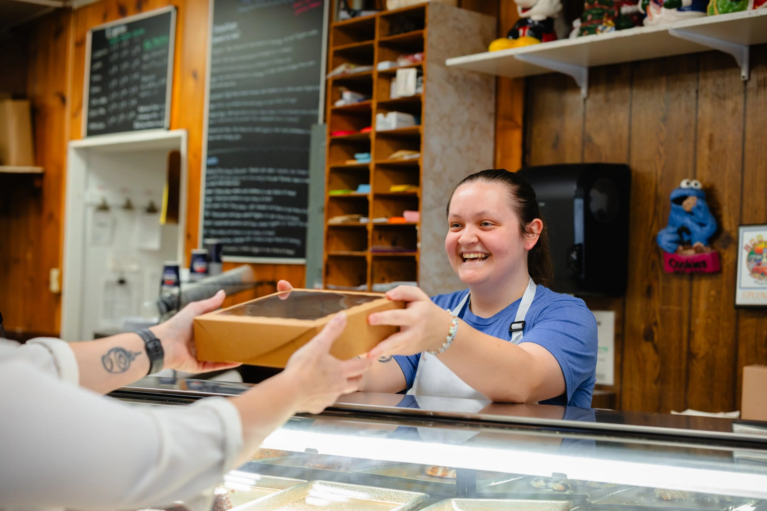 A smiling young woman in a blue shirt and apron hands a boxed cake to a customer at a bakery counter, with a wooden wall, chalkboards, and bakery items in the background.