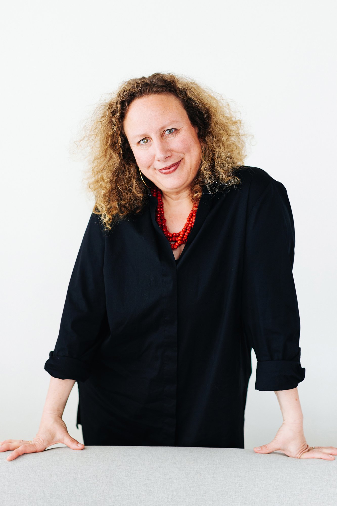 A woman with curly hair wearing a black shirt and red necklace, leaning on a table and smiling at the camera.