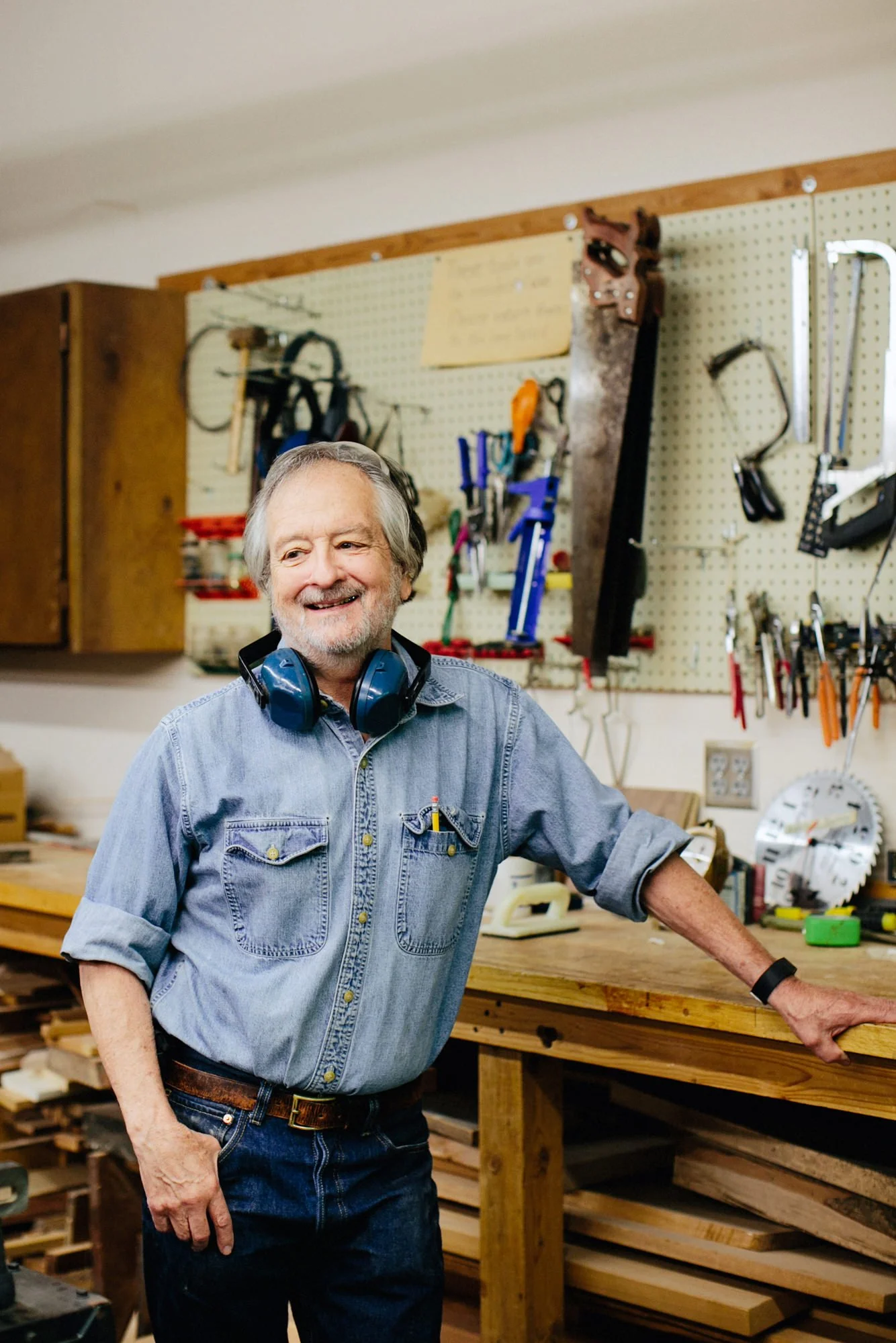 A smiling older man with gray hair and a beard, wearing a denim shirt, standing in a woodworking workshop with various tools hanging on a pegboard behind him and a wooden workbench in front.