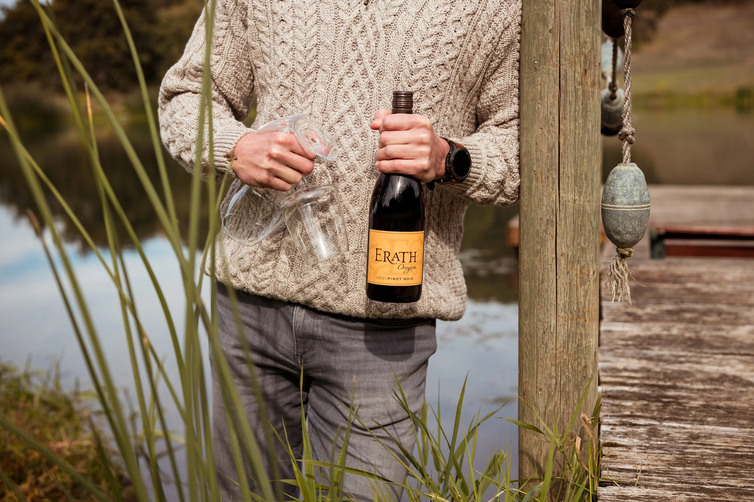 Person standing on a wooden dock near water, holding a bottle of Erath Oregon Pinot Noir and a glass, wearing a beige knit sweater and gray pants.