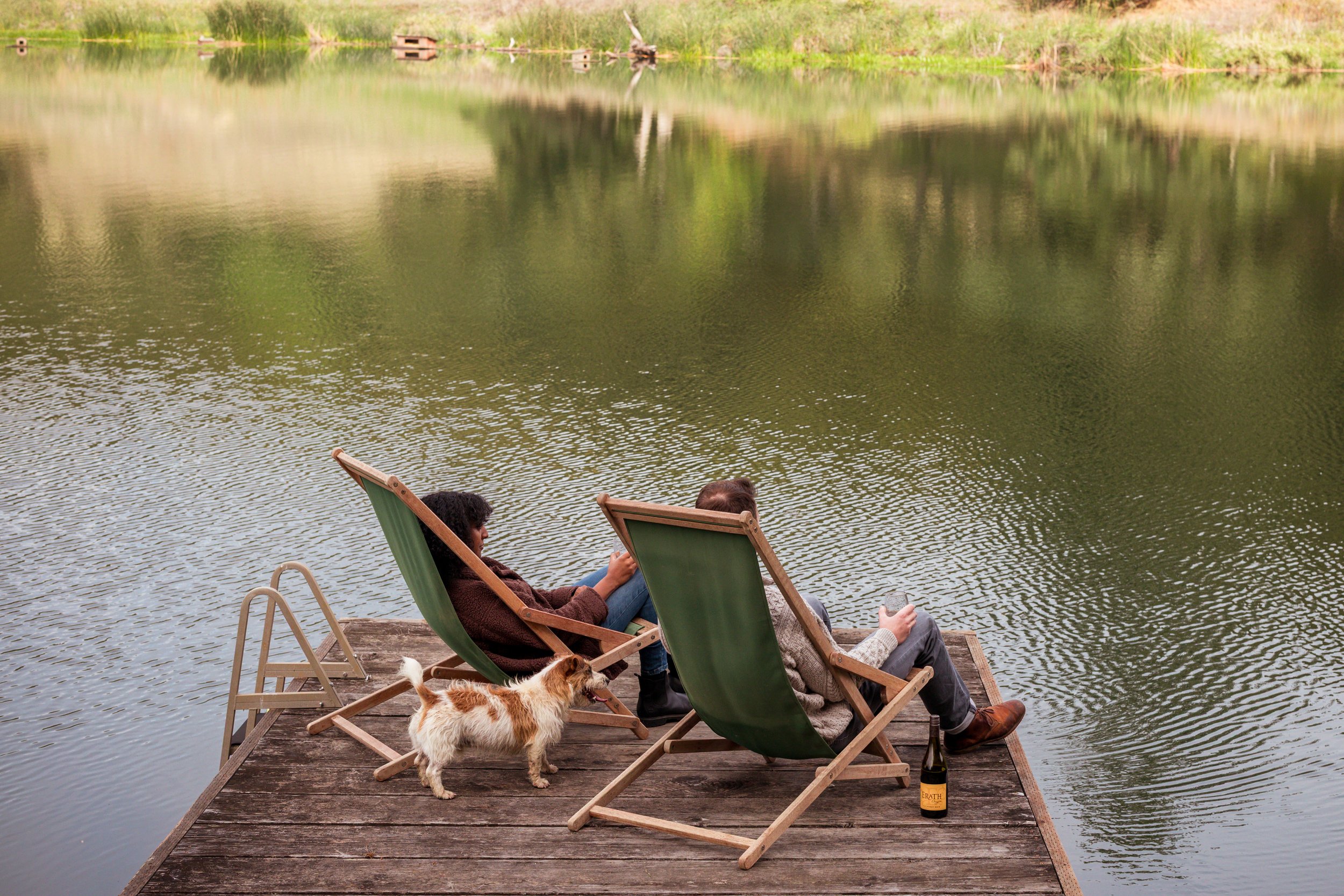 Two people relaxing on deck chairs by a lake, accompanied by a small dog. Someone is holding a smartphone and a bottle of beer is on the wooden dock.