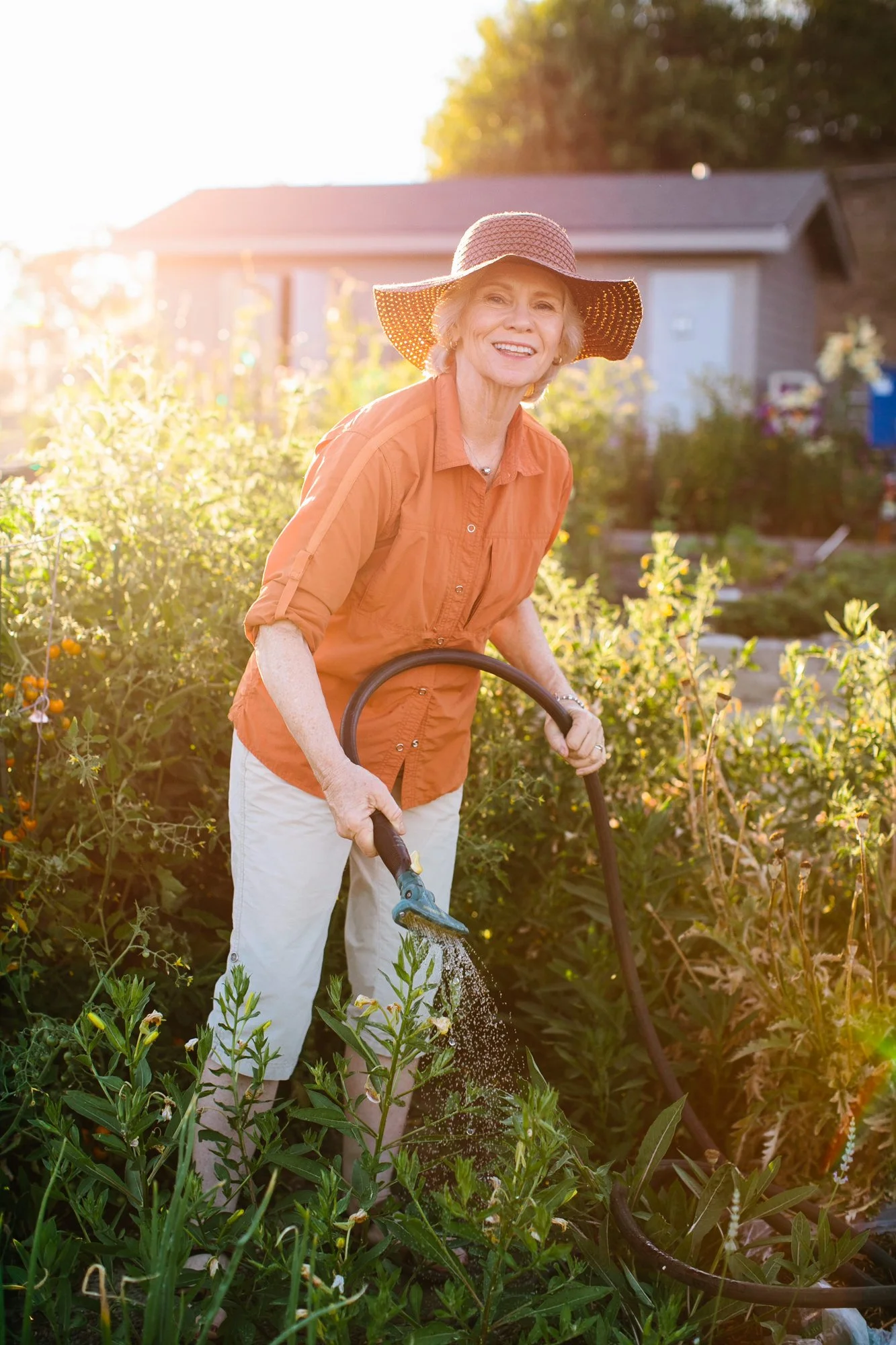 An elderly woman watering plants in her garden on a sunny day, wearing a wide-brimmed hat and an orange shirt.