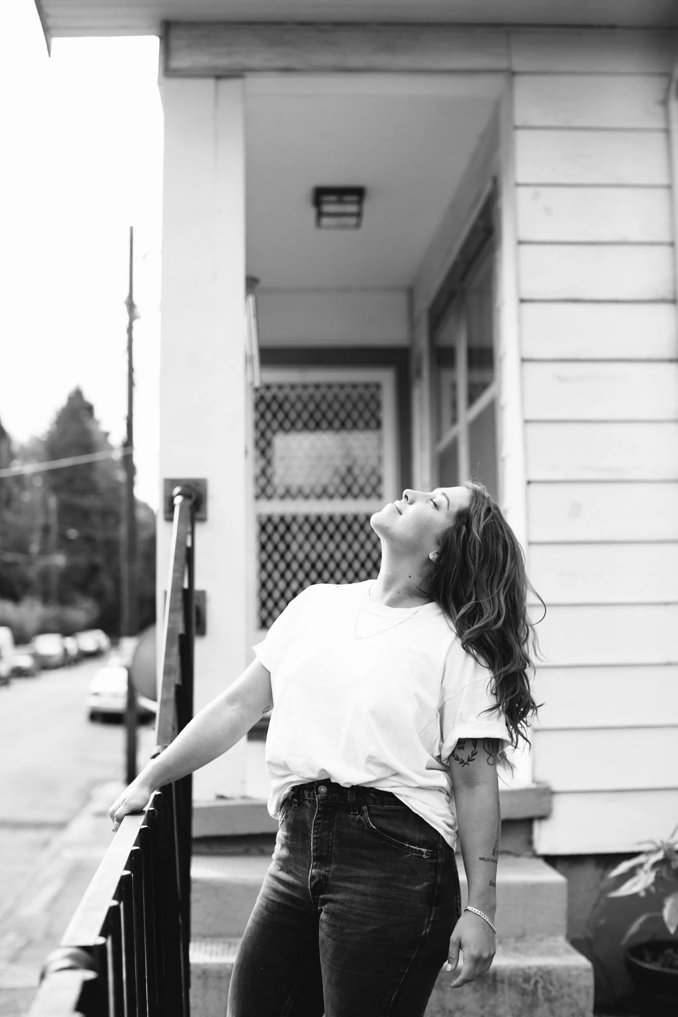 A woman with wavy hair wearing a white t-shirt and dark jeans standing outside a house, looking upward.