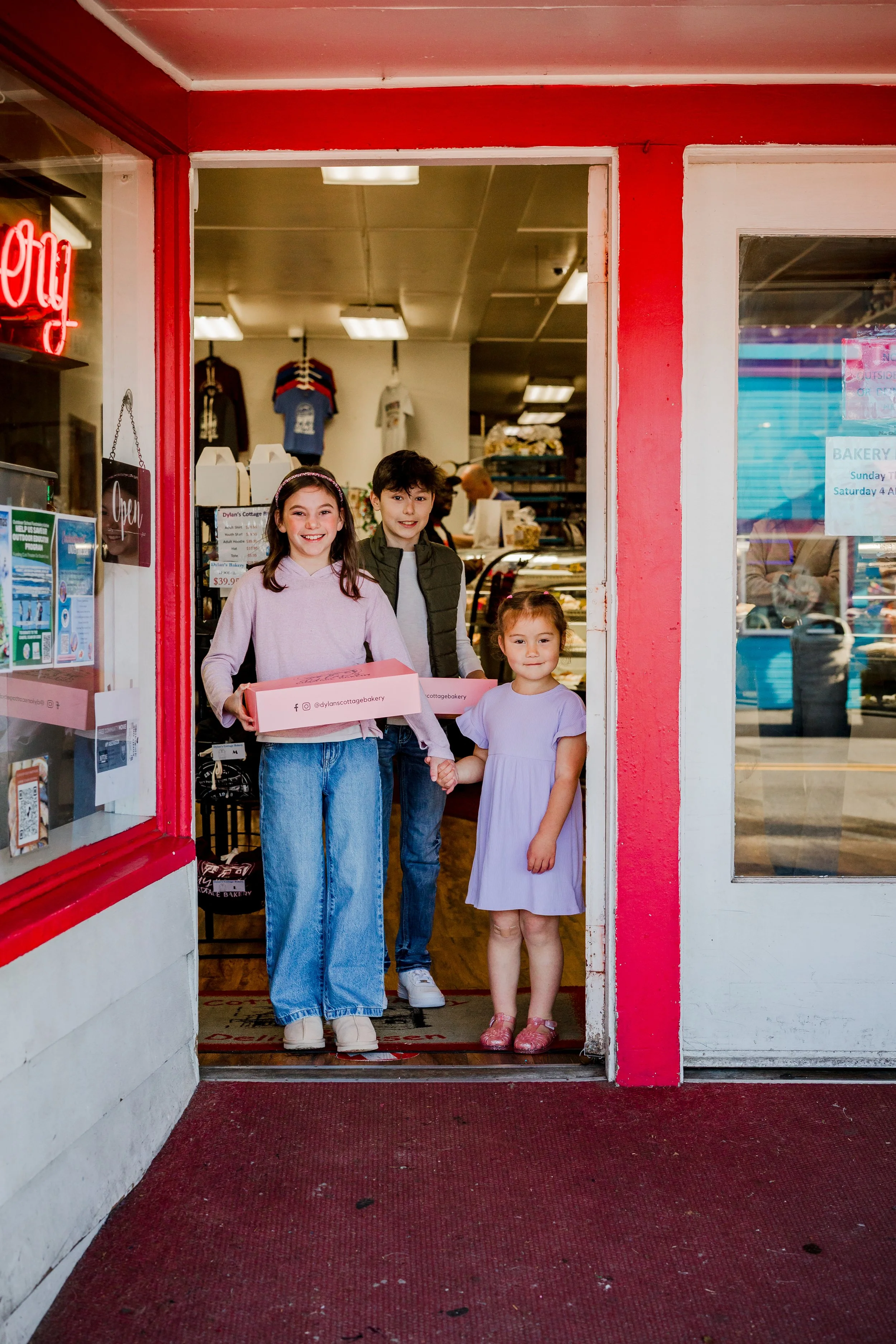 Three children standing at the entrance of a bakery, one holding a pink box, inside a store with red and white walls.