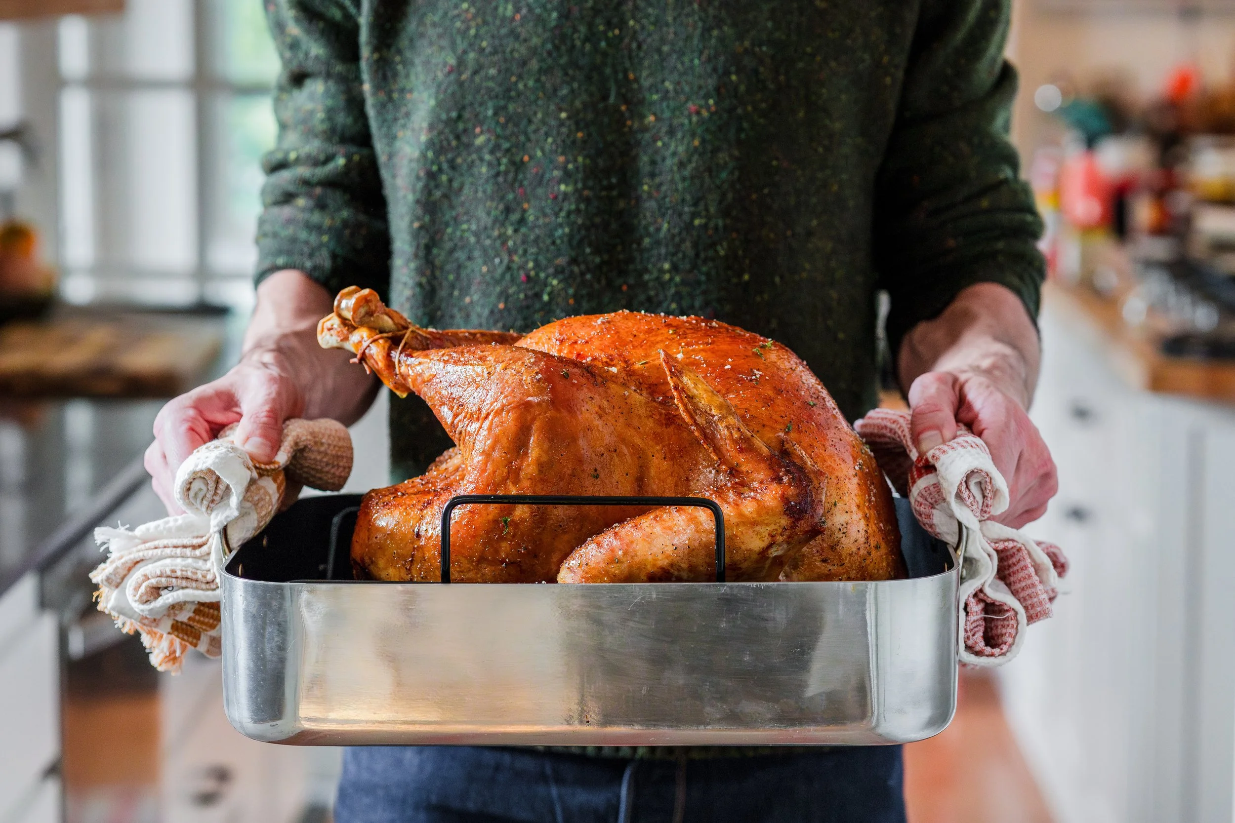 Person holding a roasted turkey in a baking pan with oven mitts inside a kitchen.
