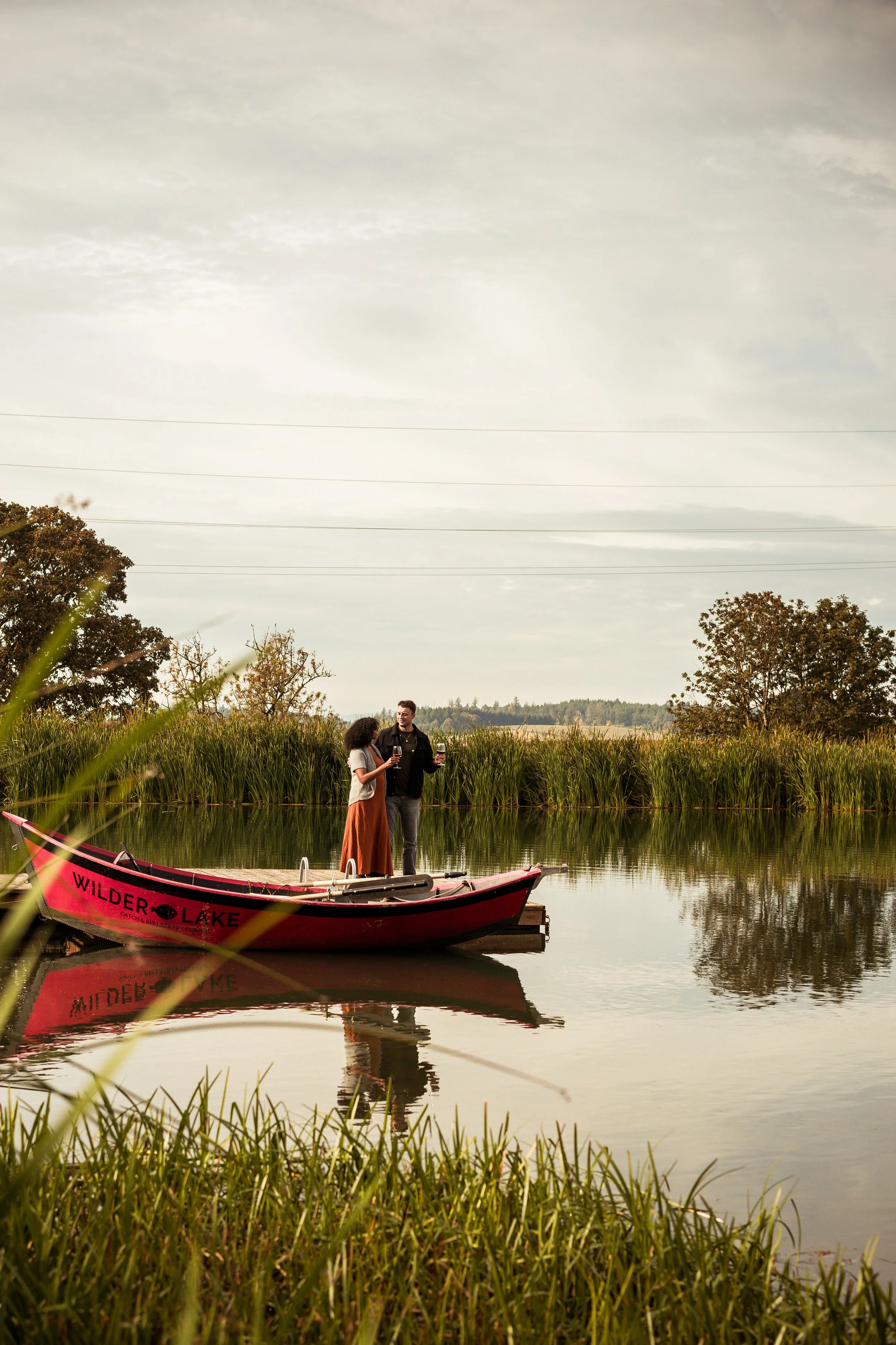 A couple standing on a dock by a lake, holding glasses of wine, with a red boat named 'Wilder Lake' floating nearby, surrounded by green grass and trees under a cloudy sky.