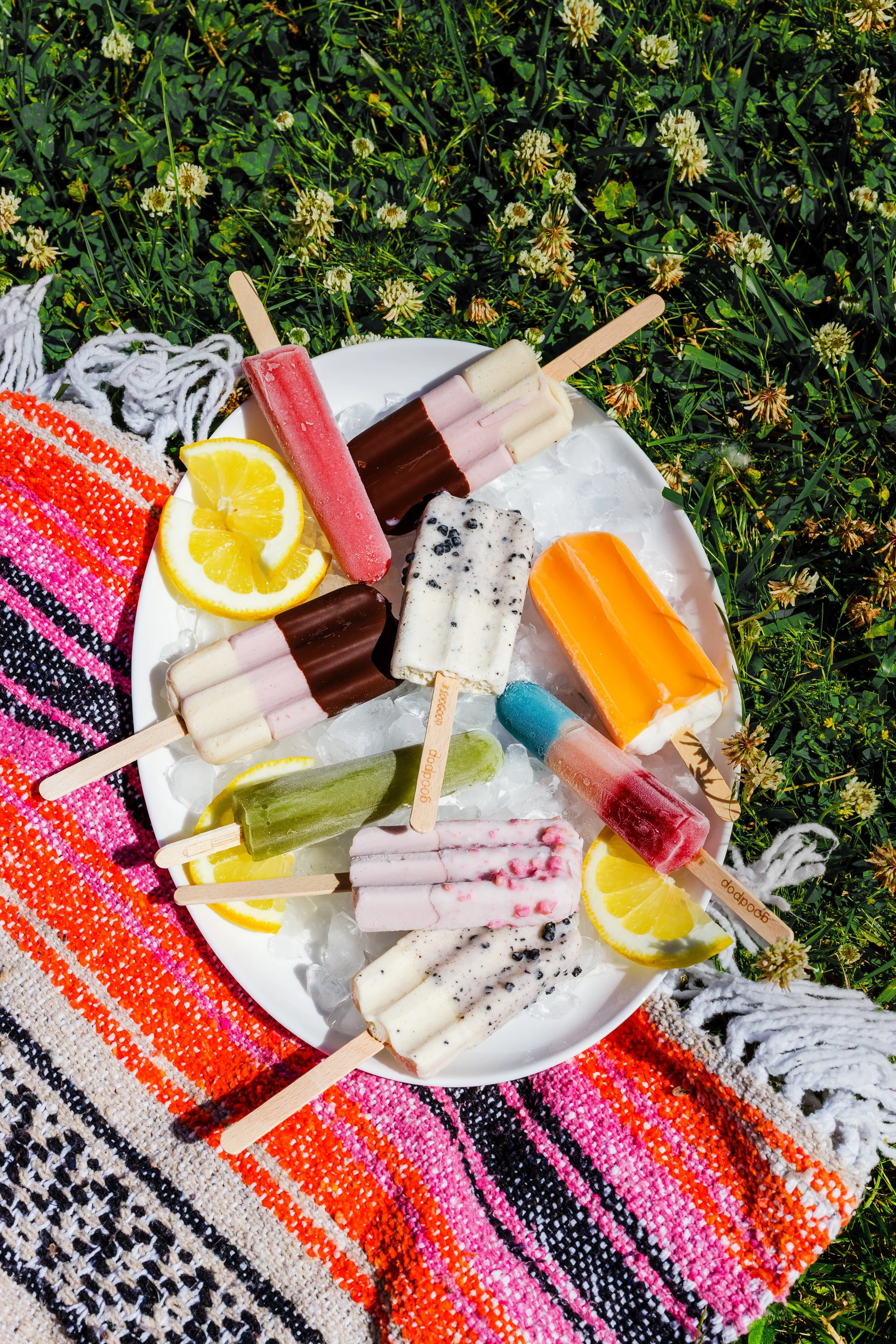 Assorted colorful ice pops on a white plate placed on a grassy surface with yellow flowers, decorated with lemon slices and a pink and orange woven cloth.