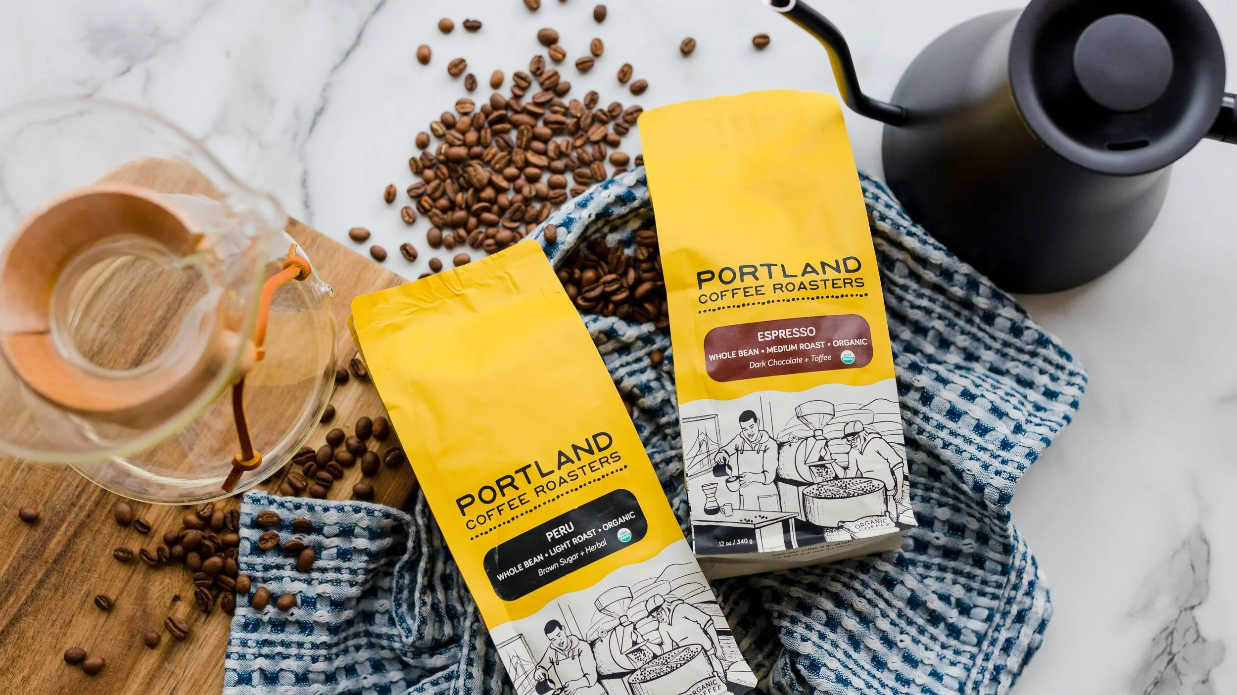 Two yellow bags of Portland Coffee Roasters coffee beans, one labeled Peru and the other labeled Espresso, are placed on a blue and white striped cloth. Coffee beans are scattered on a white marble surface and a wooden cutting board. A glass coffee server is partially visible, and a black kettle is in the background.