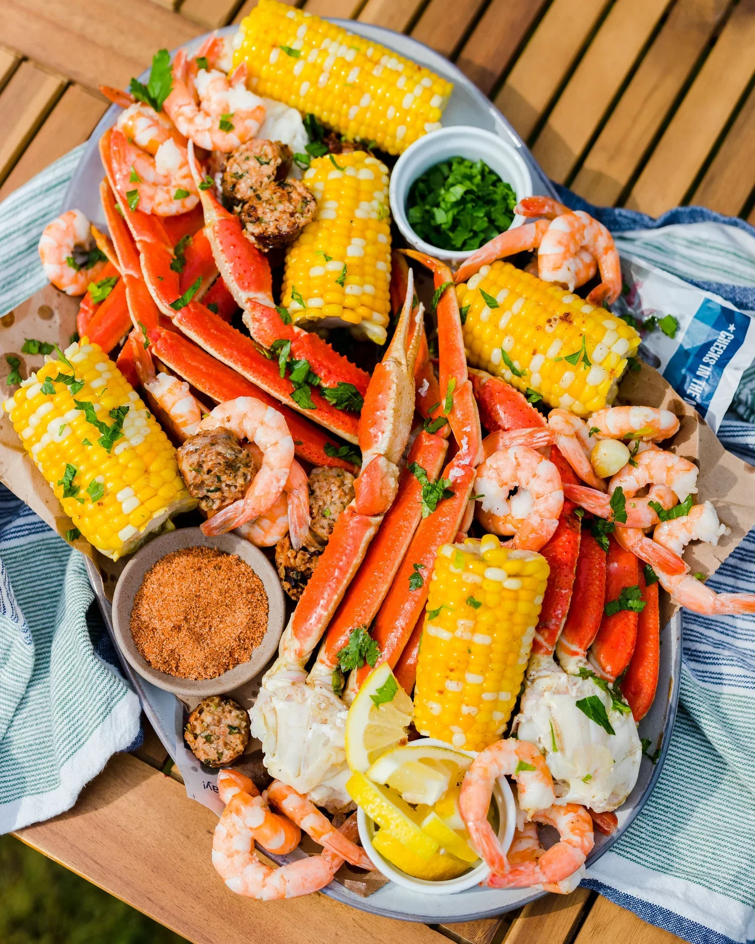 Seafood boil platter with shrimp, crab legs, corn on the cob, sausage, lemon wedges, chopped herbs, and seasoning in a small bowl.