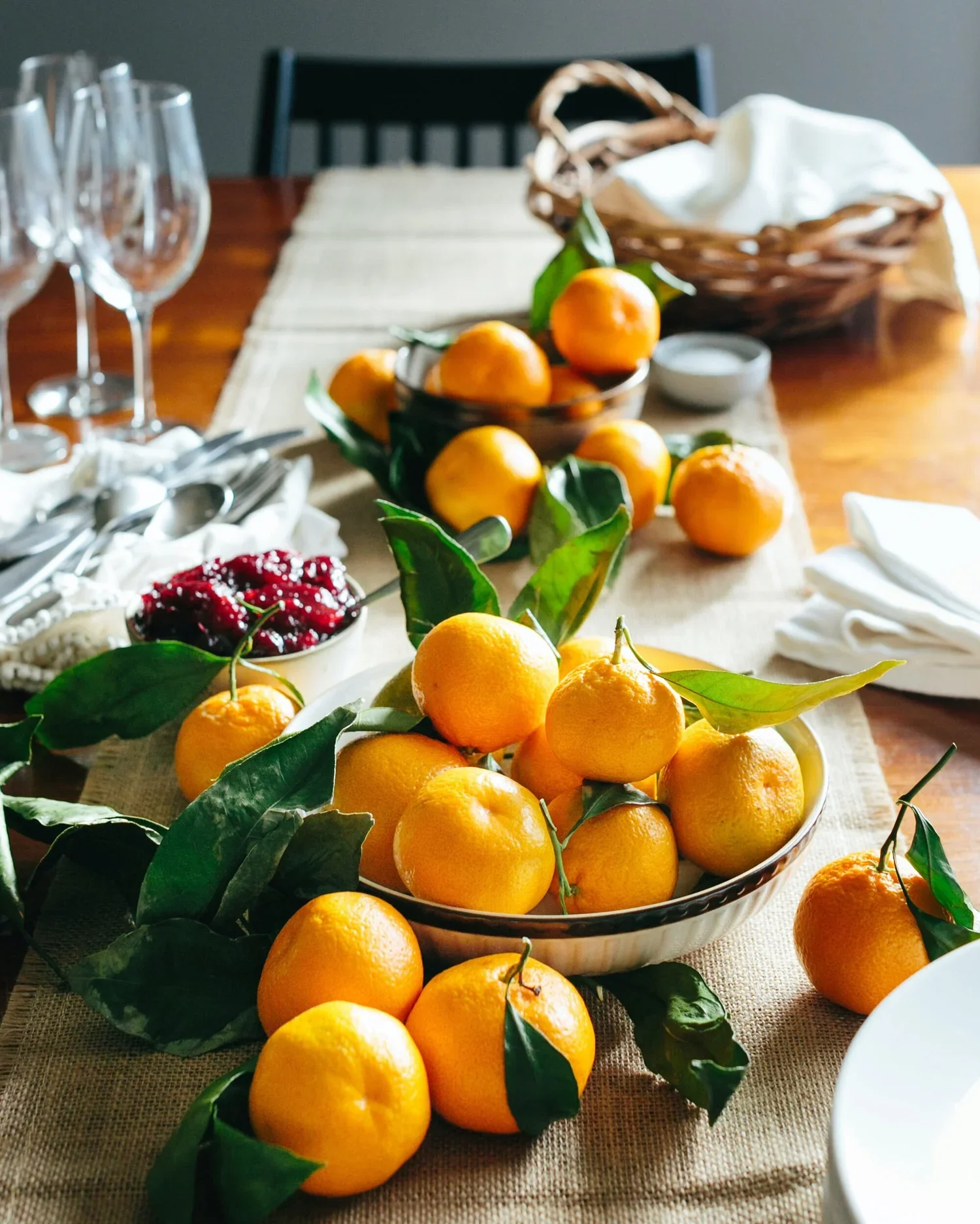 Table setting with bowls of fresh oranges and cranberries, glasses, and utensils on a table with a beige runner.