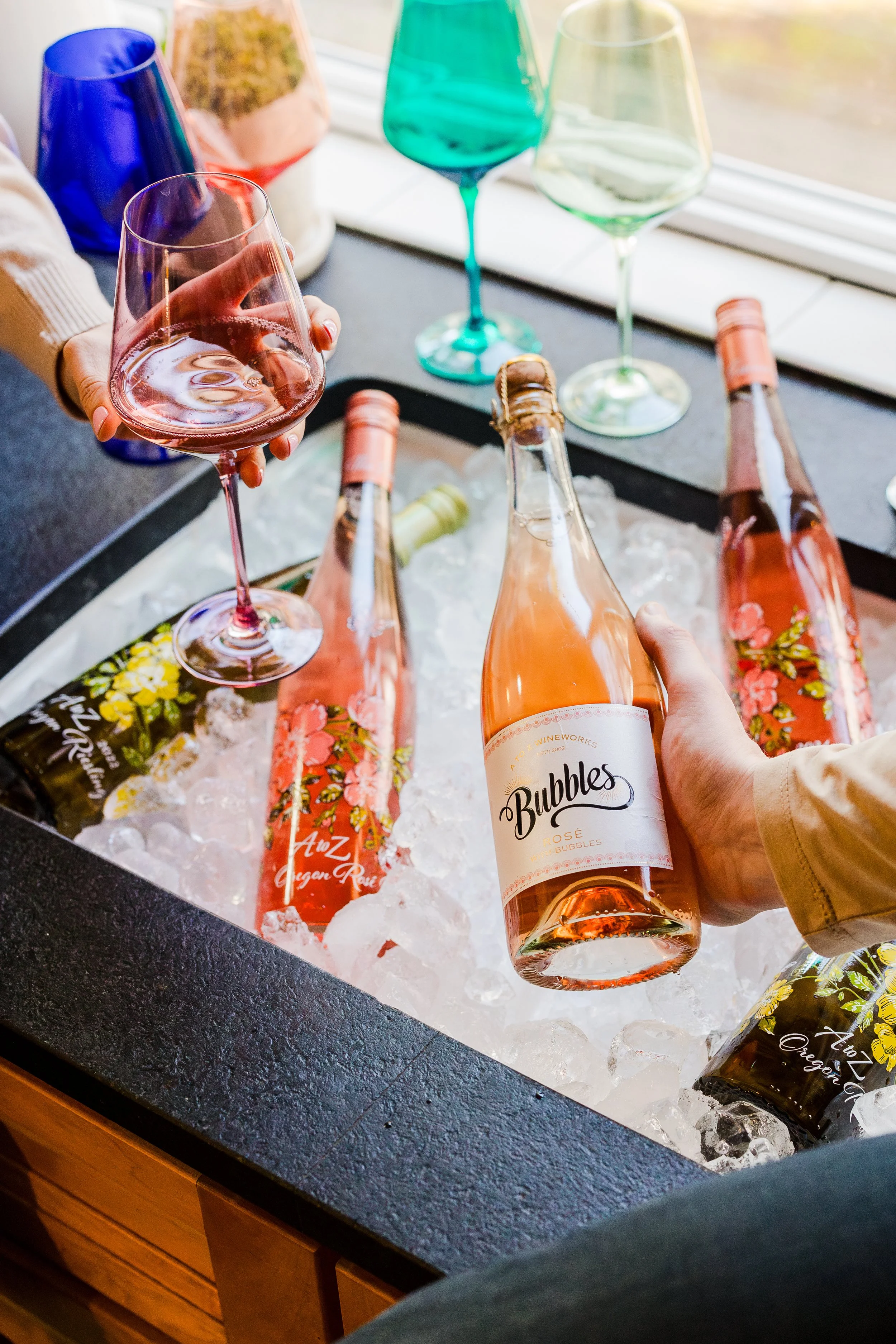 Person holding a glass of rosé wine above an ice-filled container with several bottles of rosé wine, some with floral labels, on a black countertop.
