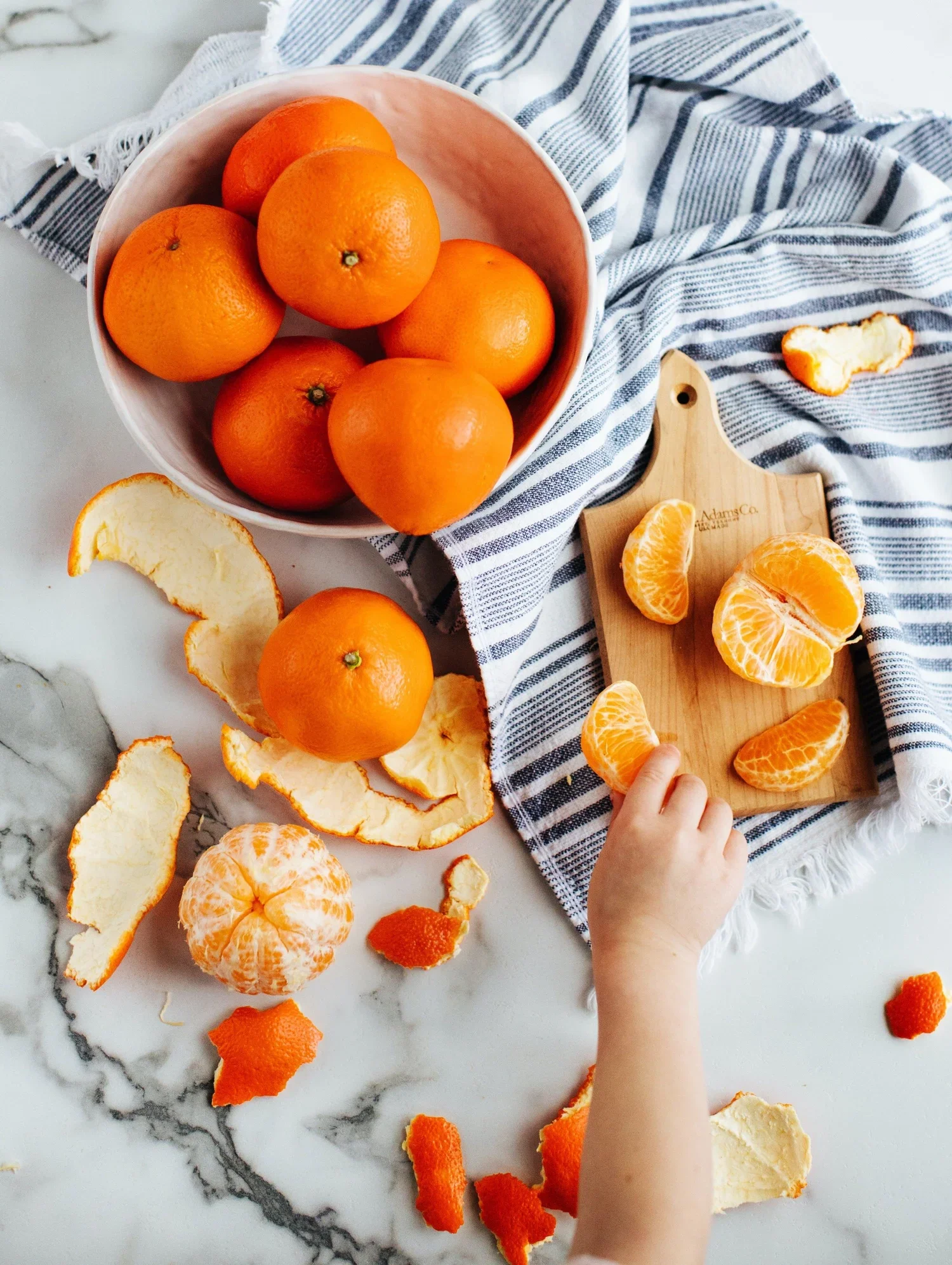 A white marble countertop with a bowl of oranges, peeled orange segments on a wooden cutting board, and some orange peel scattered around. A child's hand is reaching for a segment.