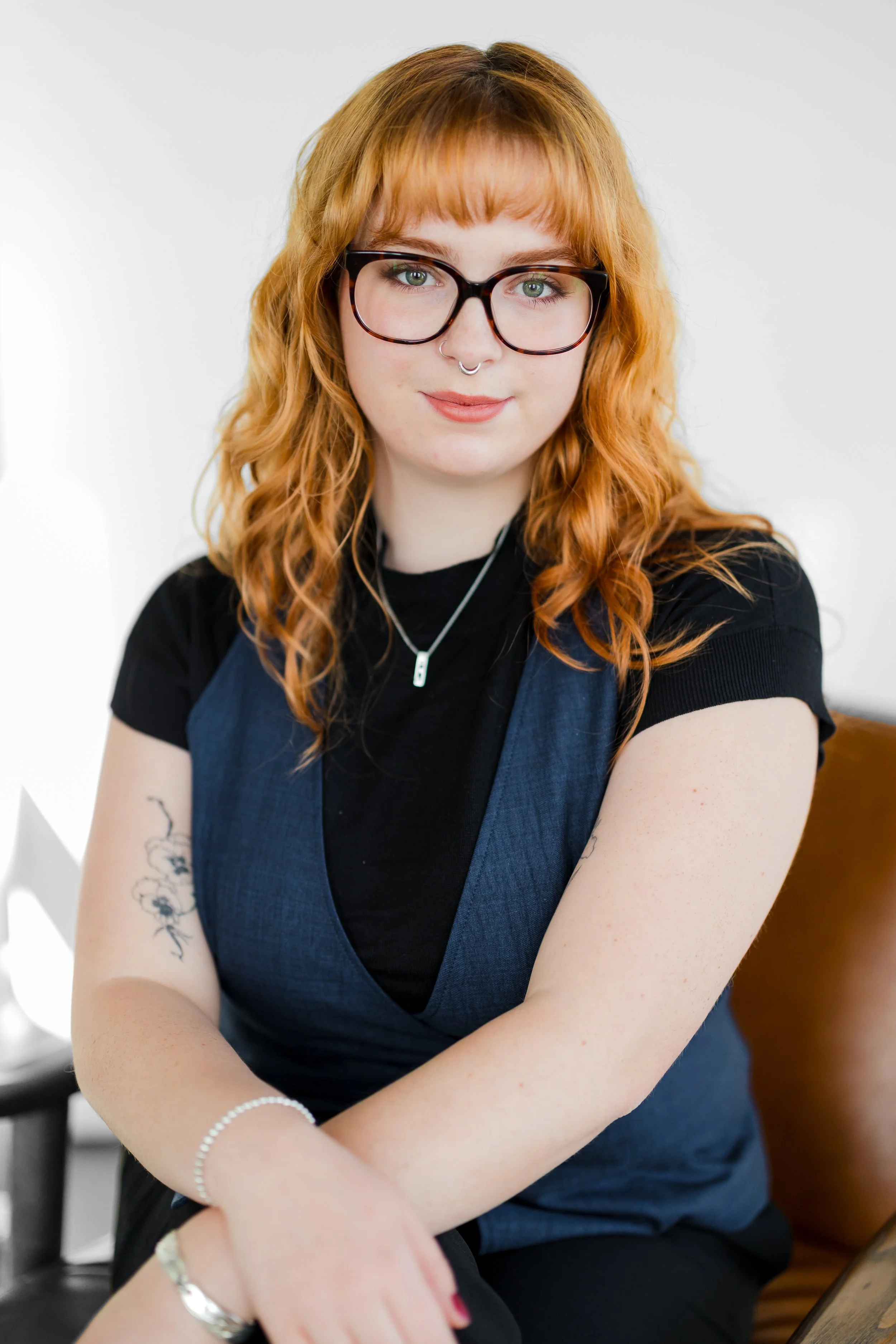 A young woman with curly red hair, glasses, and tattoos on her arm, wearing a black shirt and a blue vest, sitting on a chair with a neutral background in portland oregon