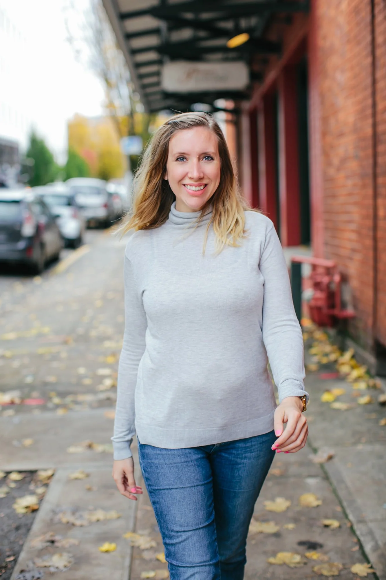 A woman with blonde hair and a light gray sweater walking on a sidewalk with fall leaves, parked cars, and a brick building in the background.